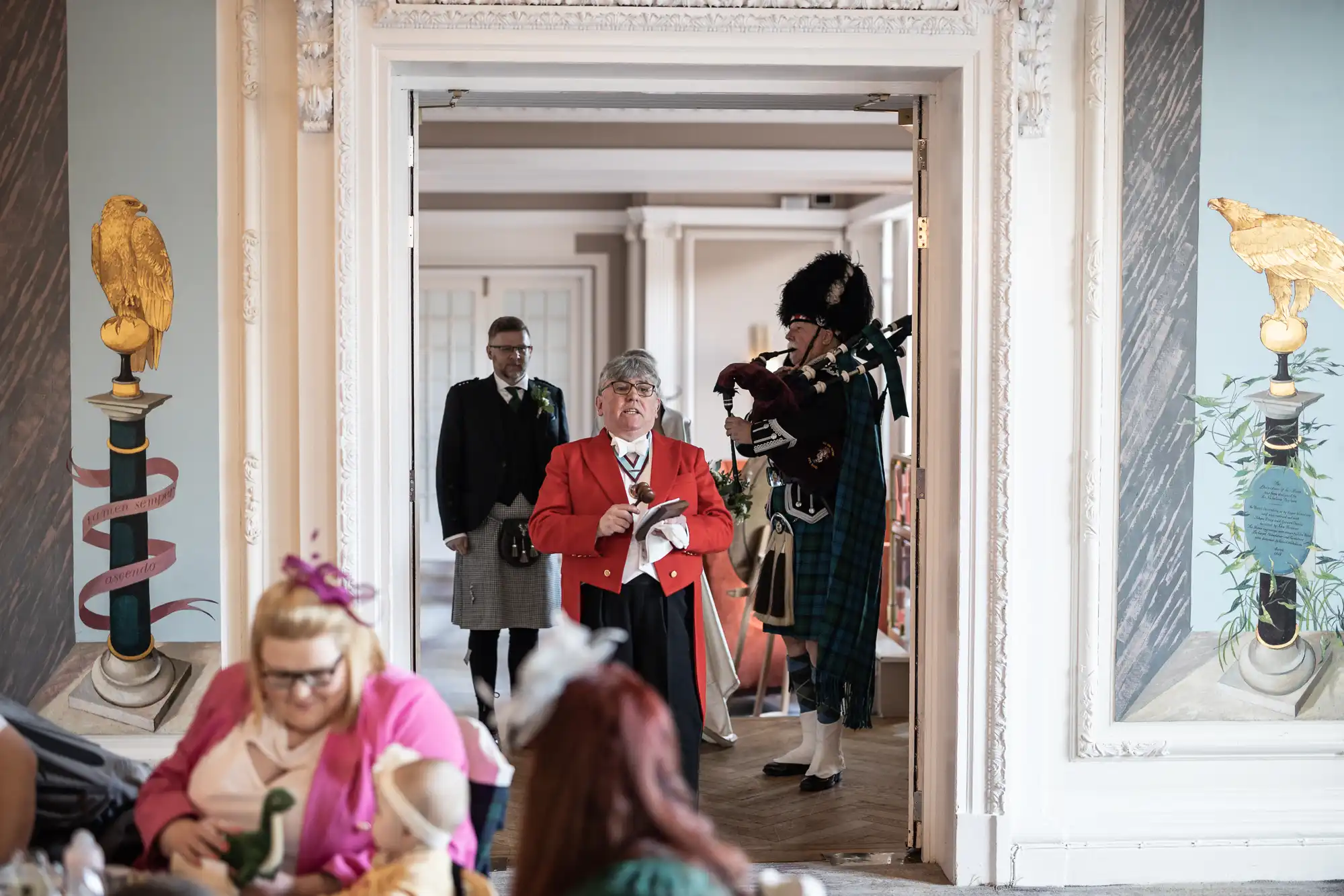 A group of people, including a piper in traditional Scottish attire, a man in a kilt, and another man in a red coat, are entering a ornately decorated room. Guests are seated at tables in the foreground.