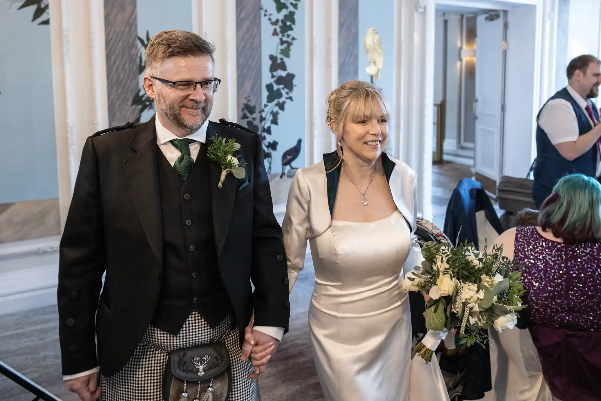 A couple, dressed in wedding attire, holds hands, smiling as they walk through a decorated venue. The man wears a dark suit with a patterned vest, and the woman wears a white gown with a green bouquet.