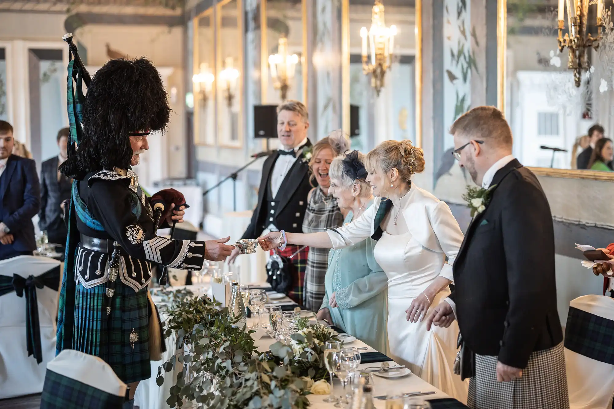 People in formal attire during a ceremonial event; a person in traditional Scottish dress hands an object to a seated woman. Decor includes greenery and elegant chandeliers.