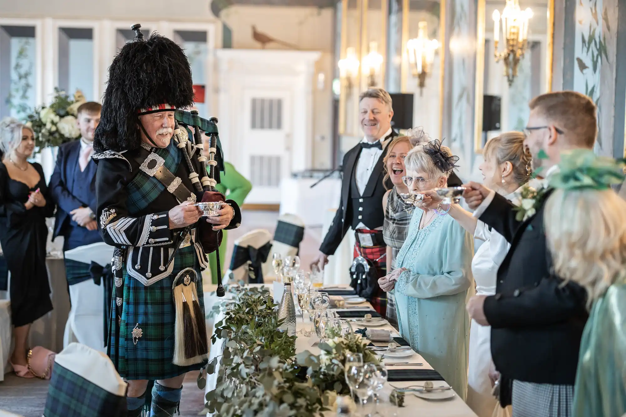 A man in traditional Scottish attire addresses guests at a formal event while others stand around a decorated table in a room with ornate lighting.