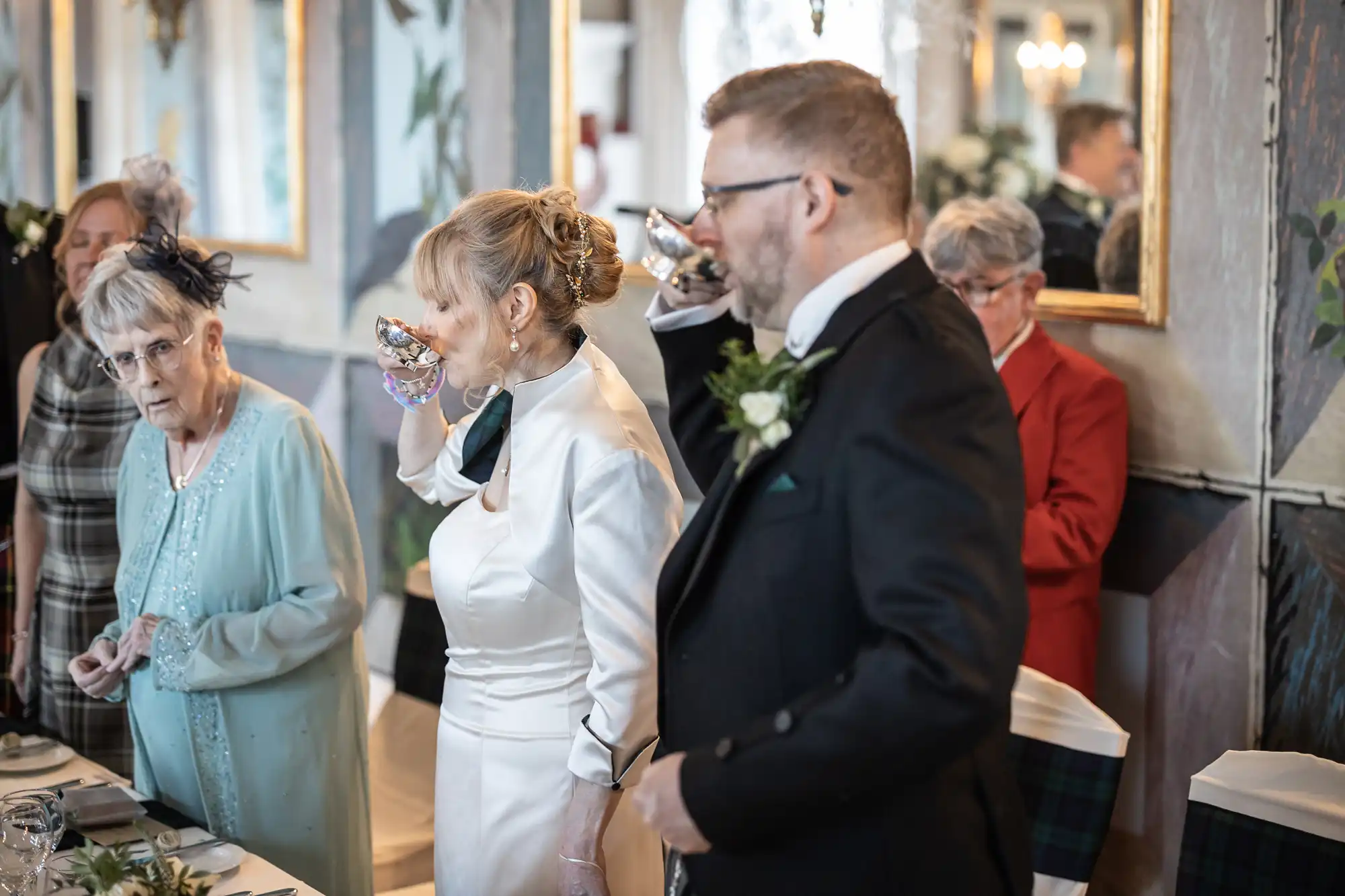 A bride in a white dress drinks from a glass, an elderly woman in blue and a man in a suit beside her, at a dining table with other people in the background.