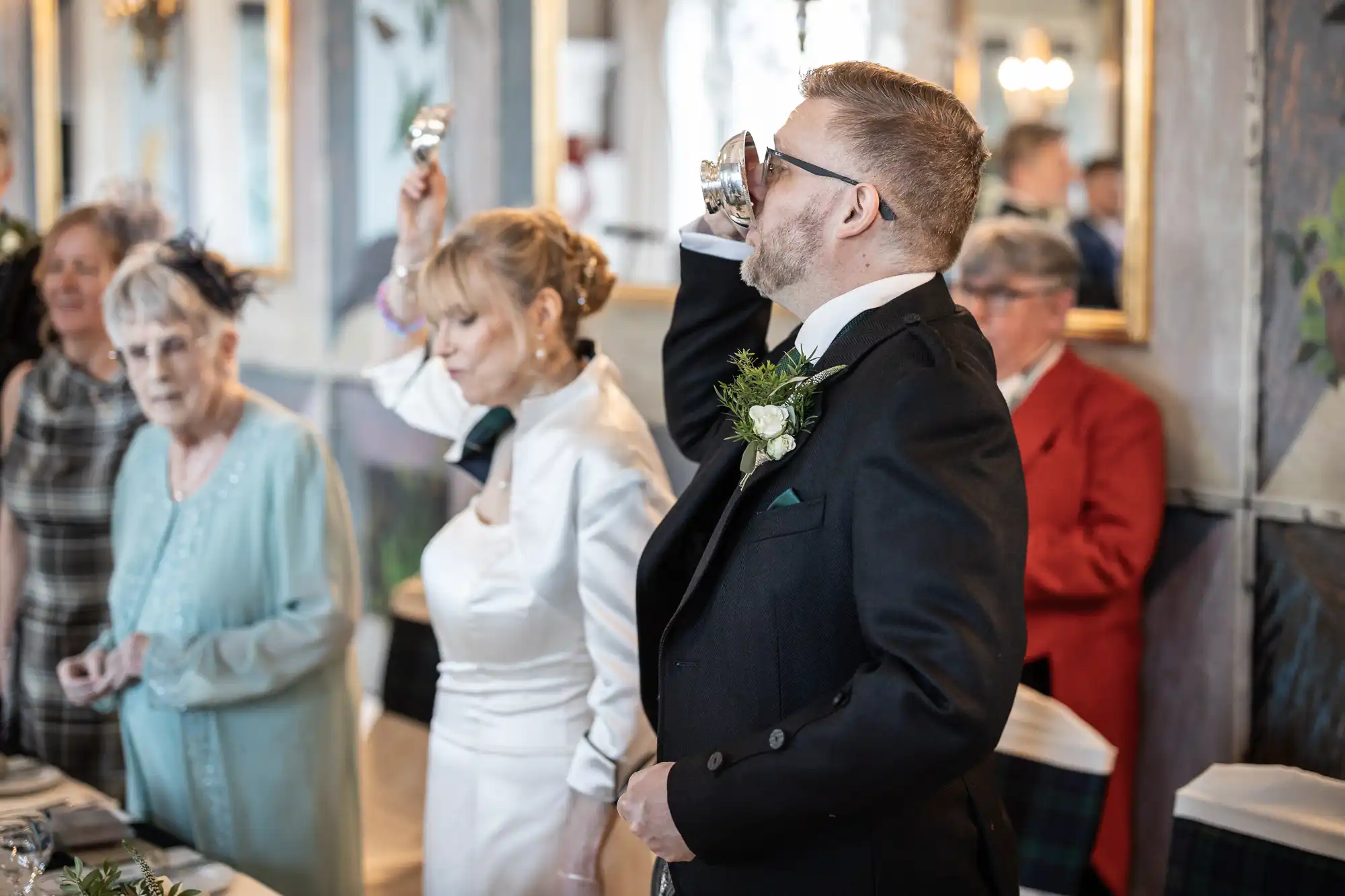 A man in formal attire drinks from a cup at a gathering. An elegantly dressed woman stands beside him, holding a cup in the air. Other attendees are seen in the background.