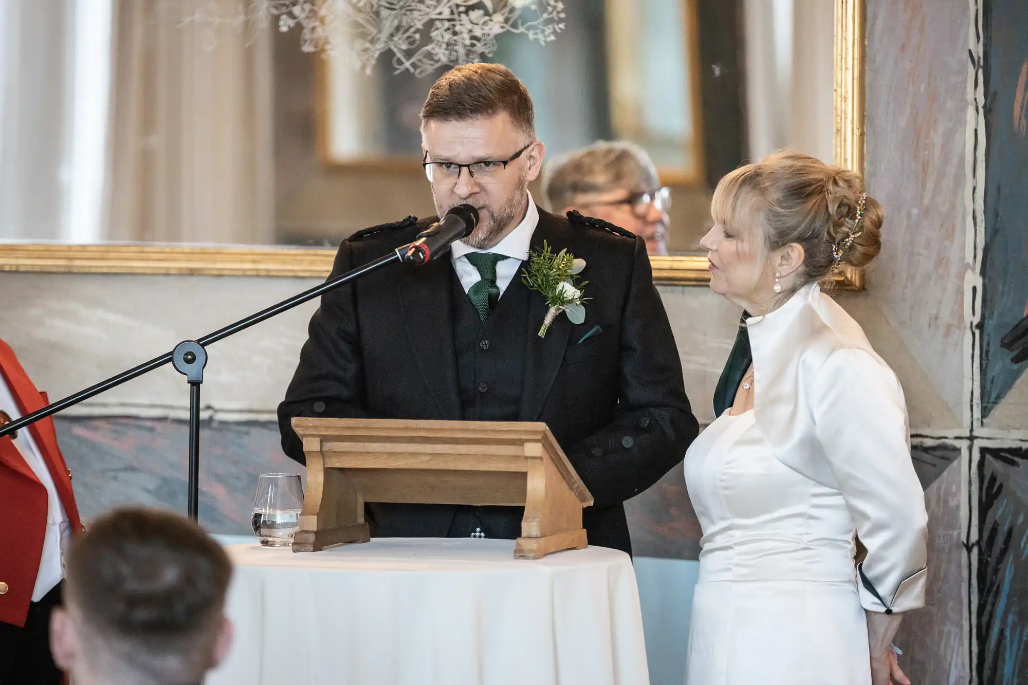 A man in formal Scottish attire speaks into a microphone at a podium next to a woman in a white dress during an indoor event.