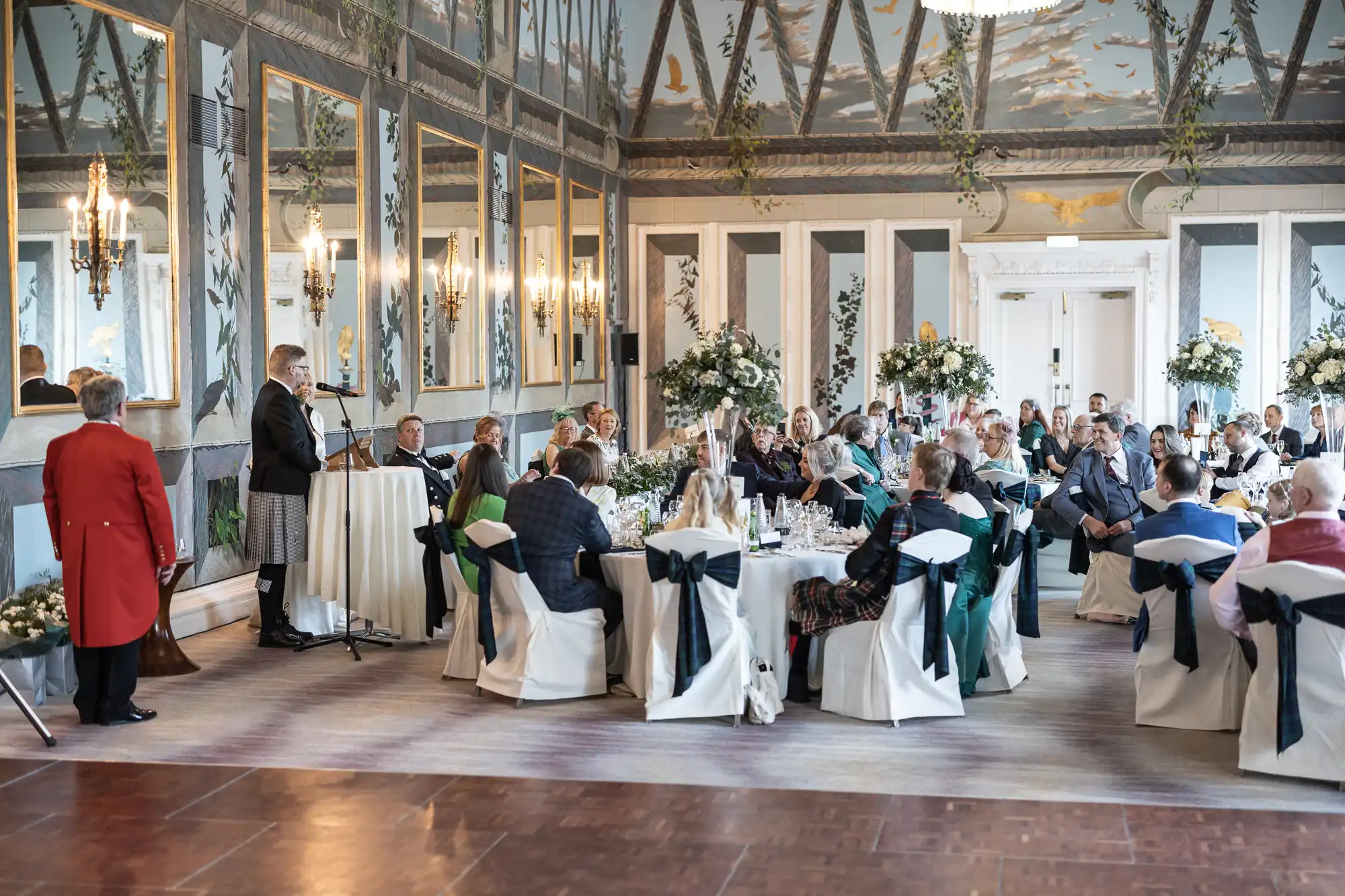 A formal event with attendees seated at round tables in an elegant, ornately decorated room with large mirrors and chandeliers. A speaker dressed in a suit stands at a podium.