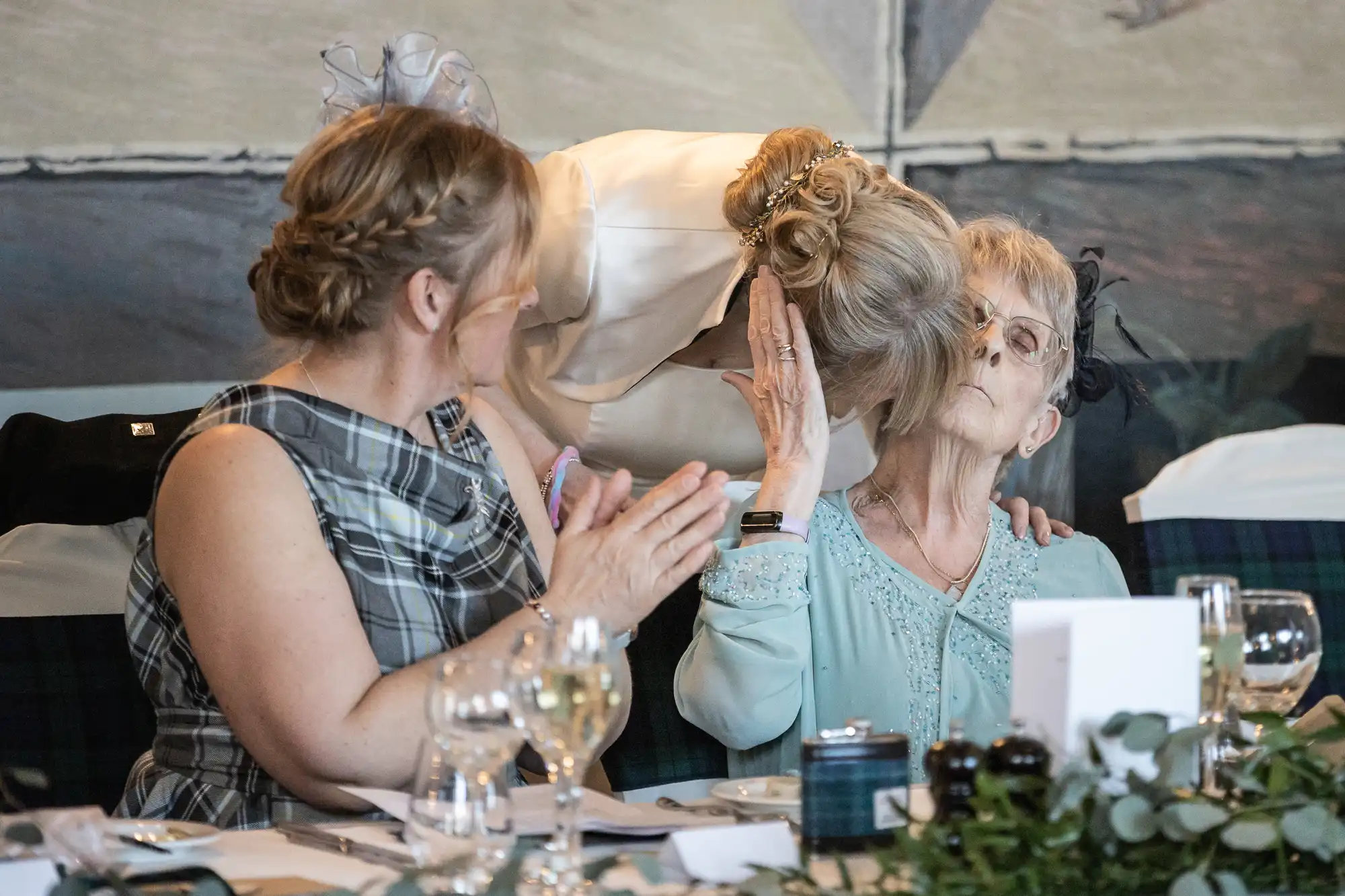 A bride leans in to kiss an elderly woman on the cheek while another woman in a plaid dress sits beside them, clapping.