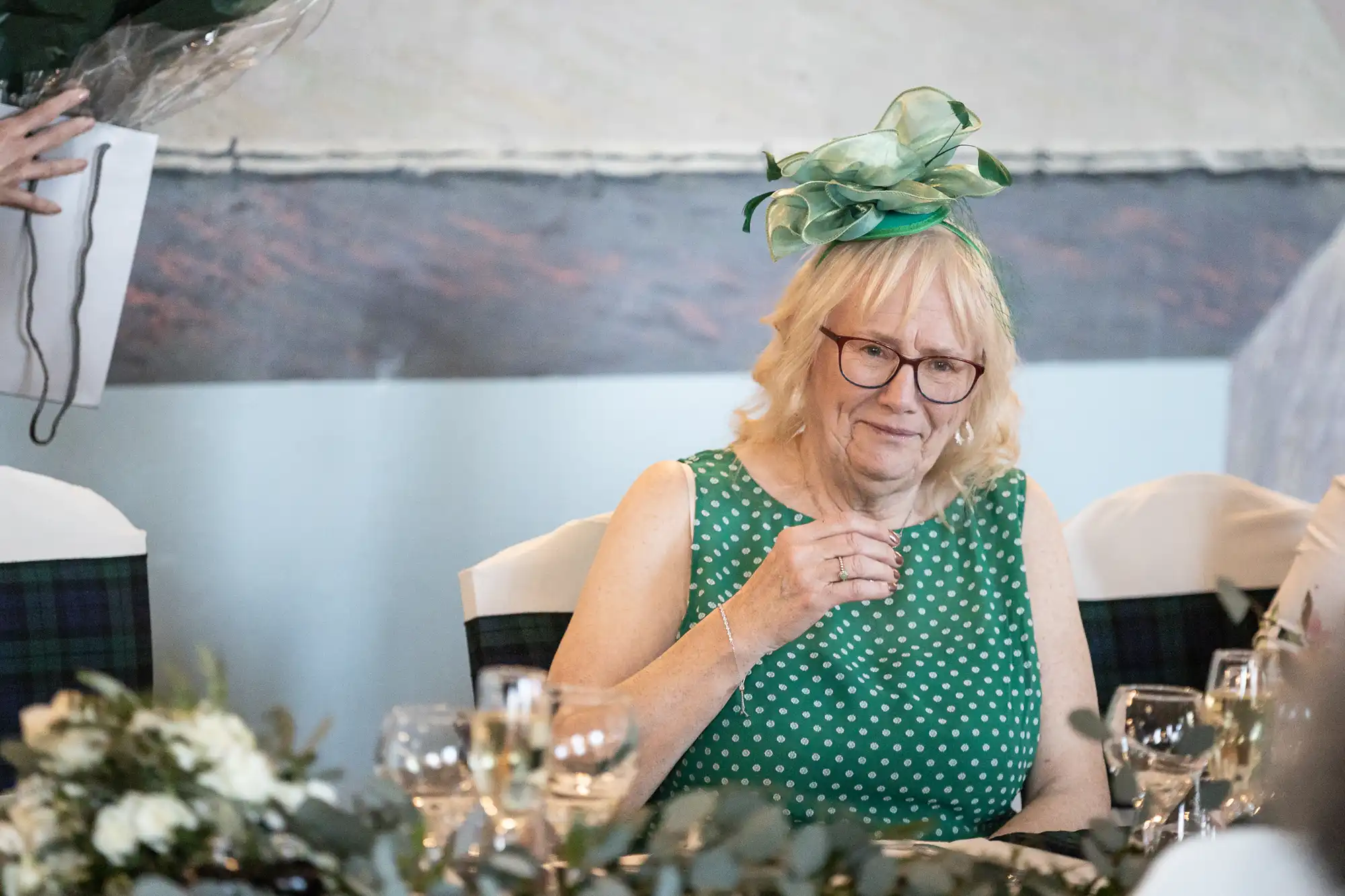 An elderly woman wearing glasses and a green dress with white polka dots sits at a table holding a drink, with a flower arrangement and multiple wine glasses in front of her. She wears a green fascinator.