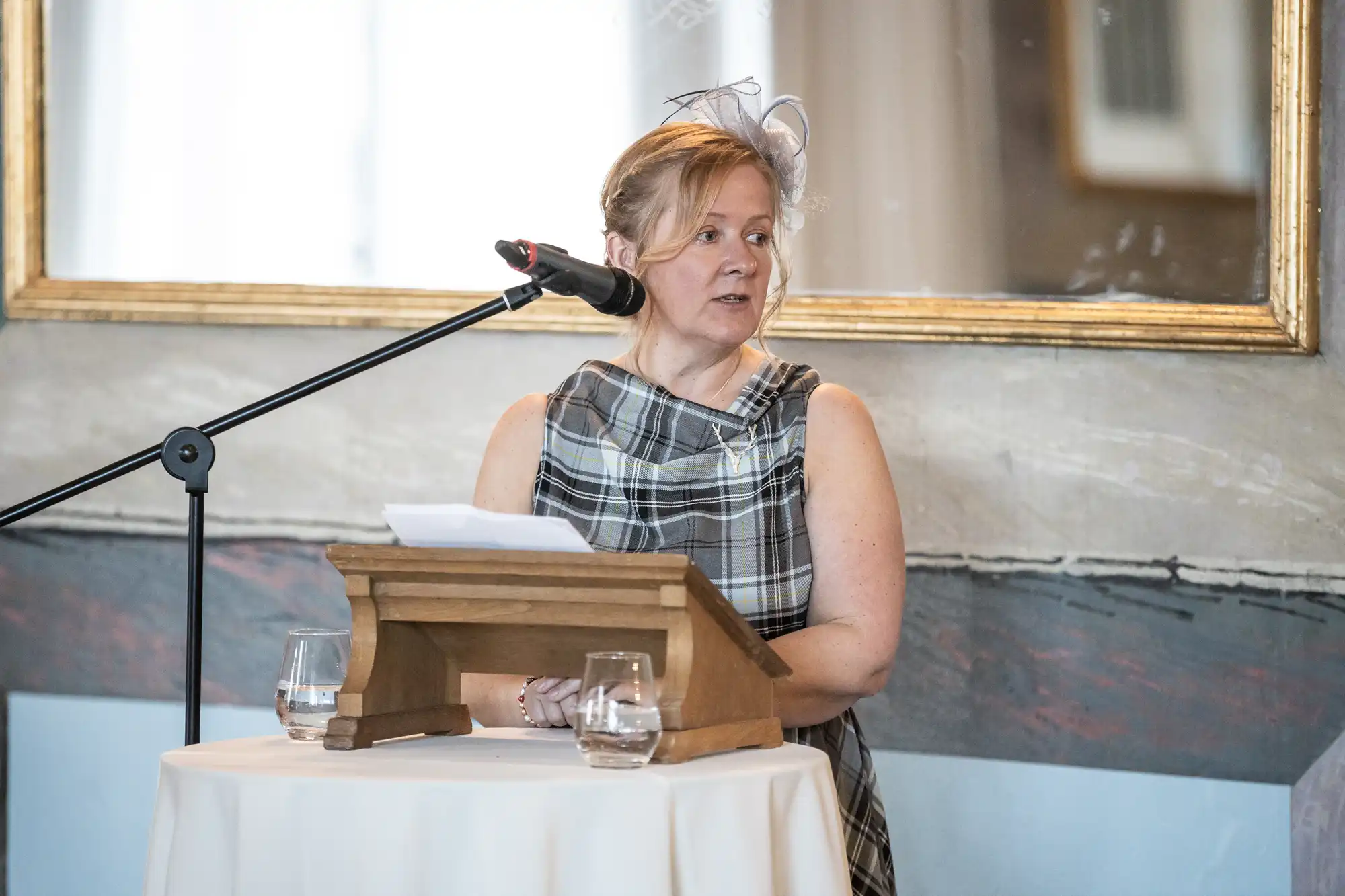 A woman stands at a podium with a microphone and reads from her notes. She is wearing a plaid dress and a small fascinator hat. Two glasses of water are on the round table beside her.