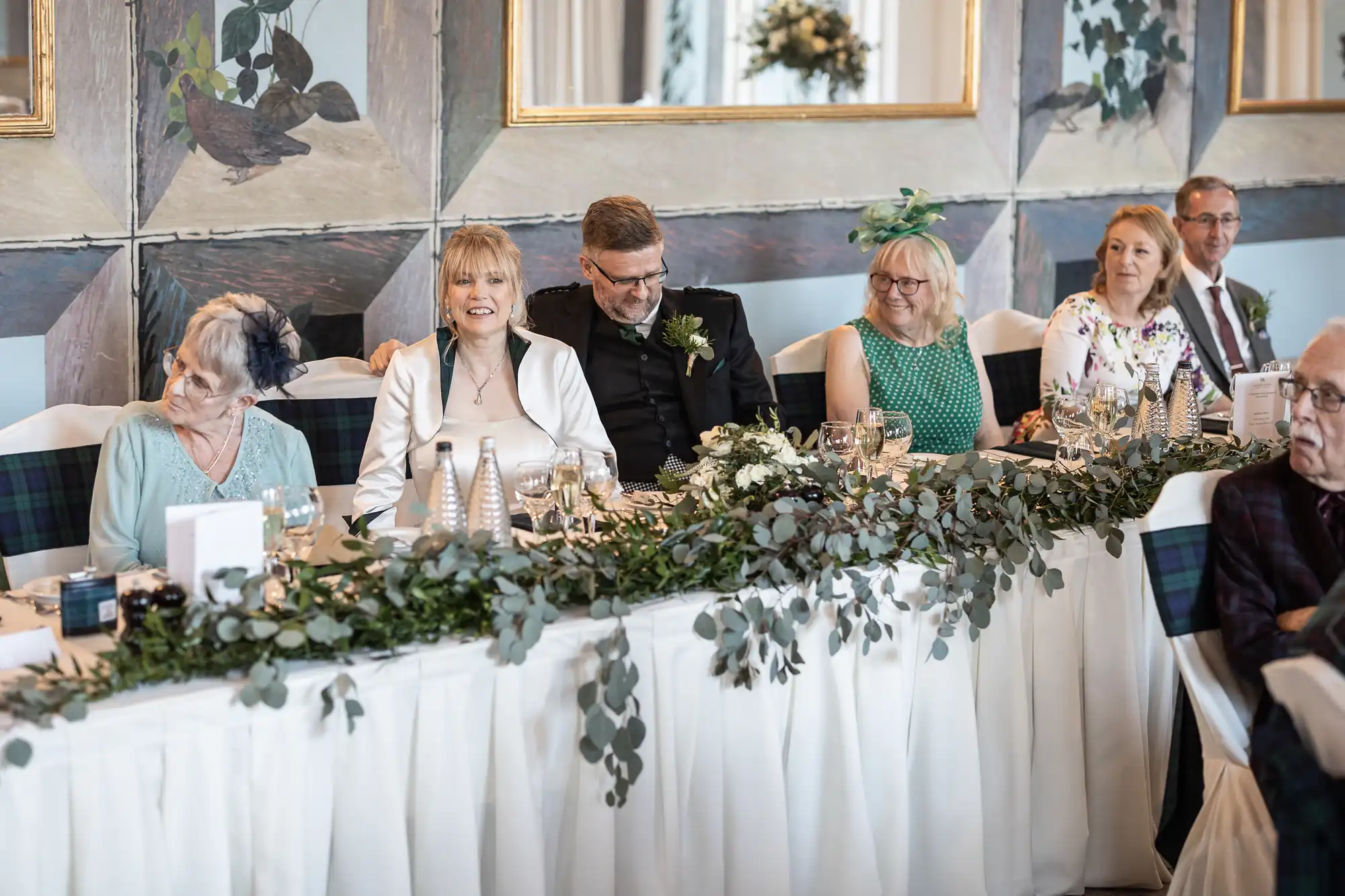 A group of people, including an older and a younger couple, sit at a long table adorned with greenery and tableware in a formal setting, possibly during a wedding reception or formal event.