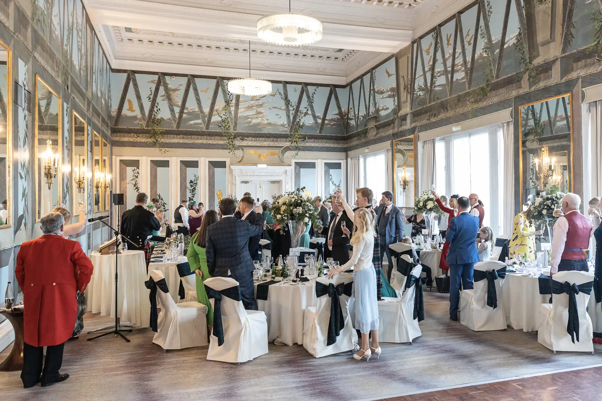 Guests dressed in formal attire raise glasses for a toast in an elegantly decorated banquet hall with large windows and wall mirrors.