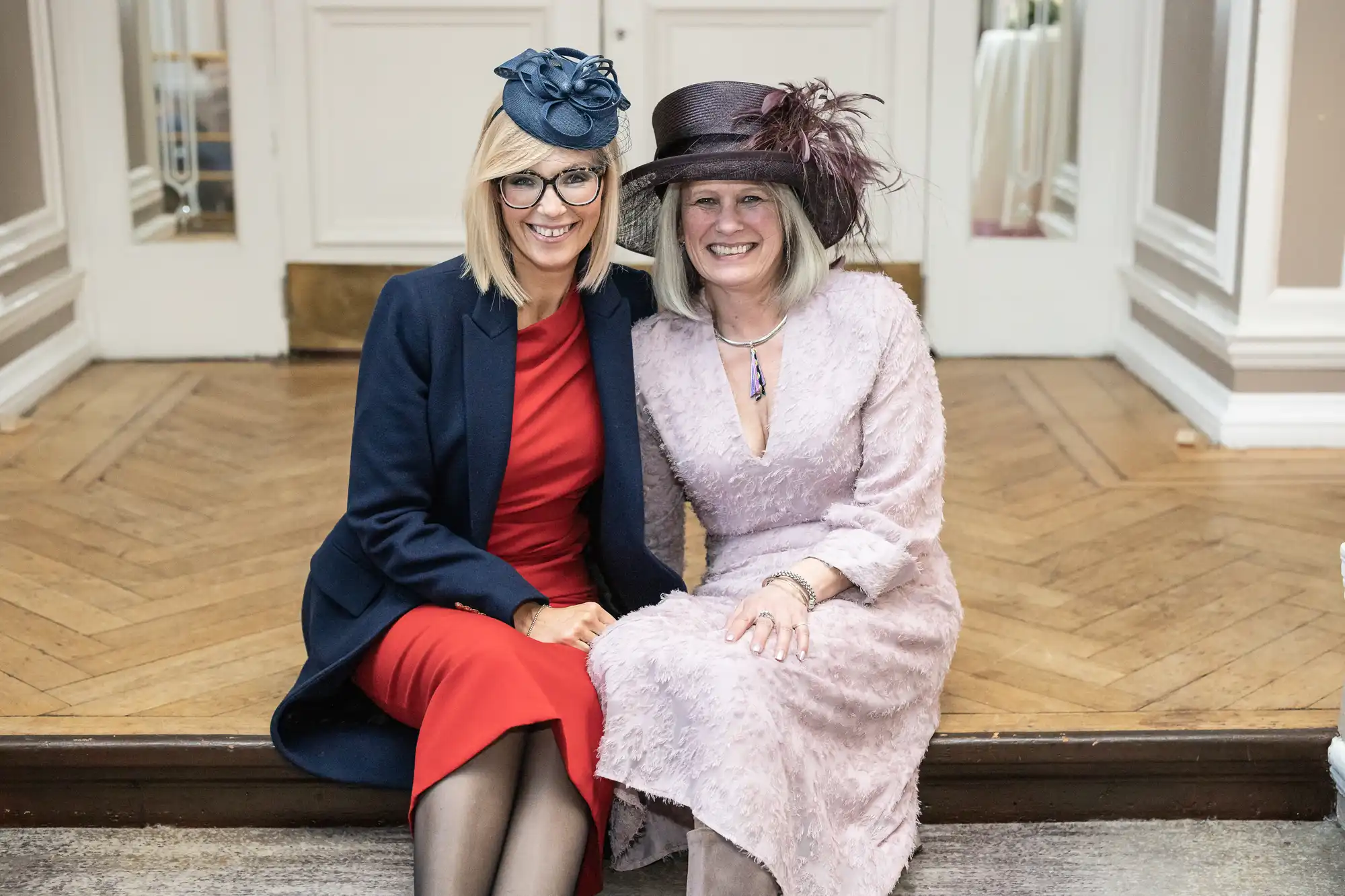 Two women, dressed formally with hats, sit side by side on a step, smiling at the camera. One wears a red dress with a dark blazer, the other wears a light pink dress with a brown hat.