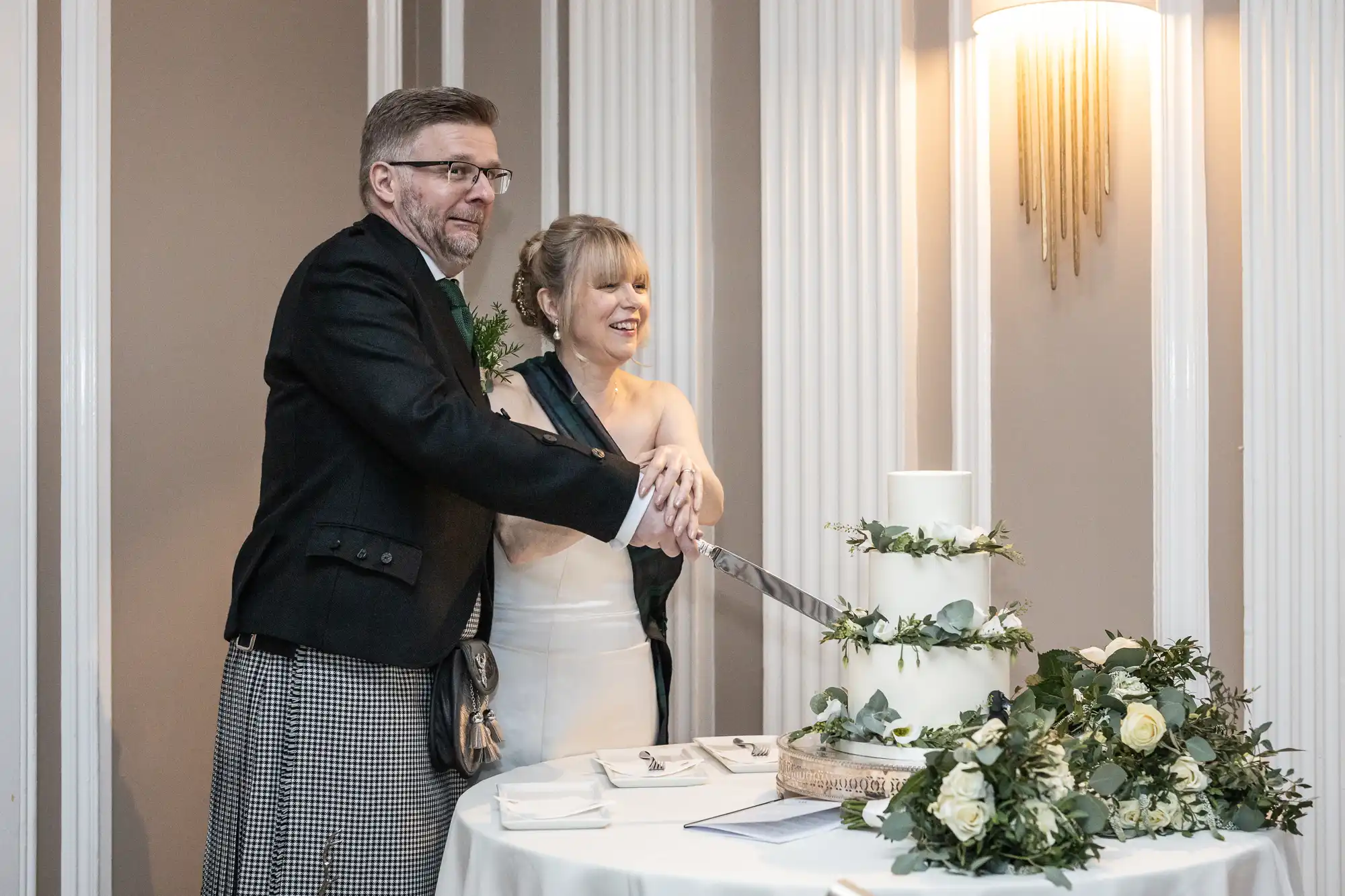 A couple, dressed in formal attire, cuts a white, multi-tiered cake adorned with green and white floral decorations, standing at a round table in a well-lit room with white vertical wall panels.