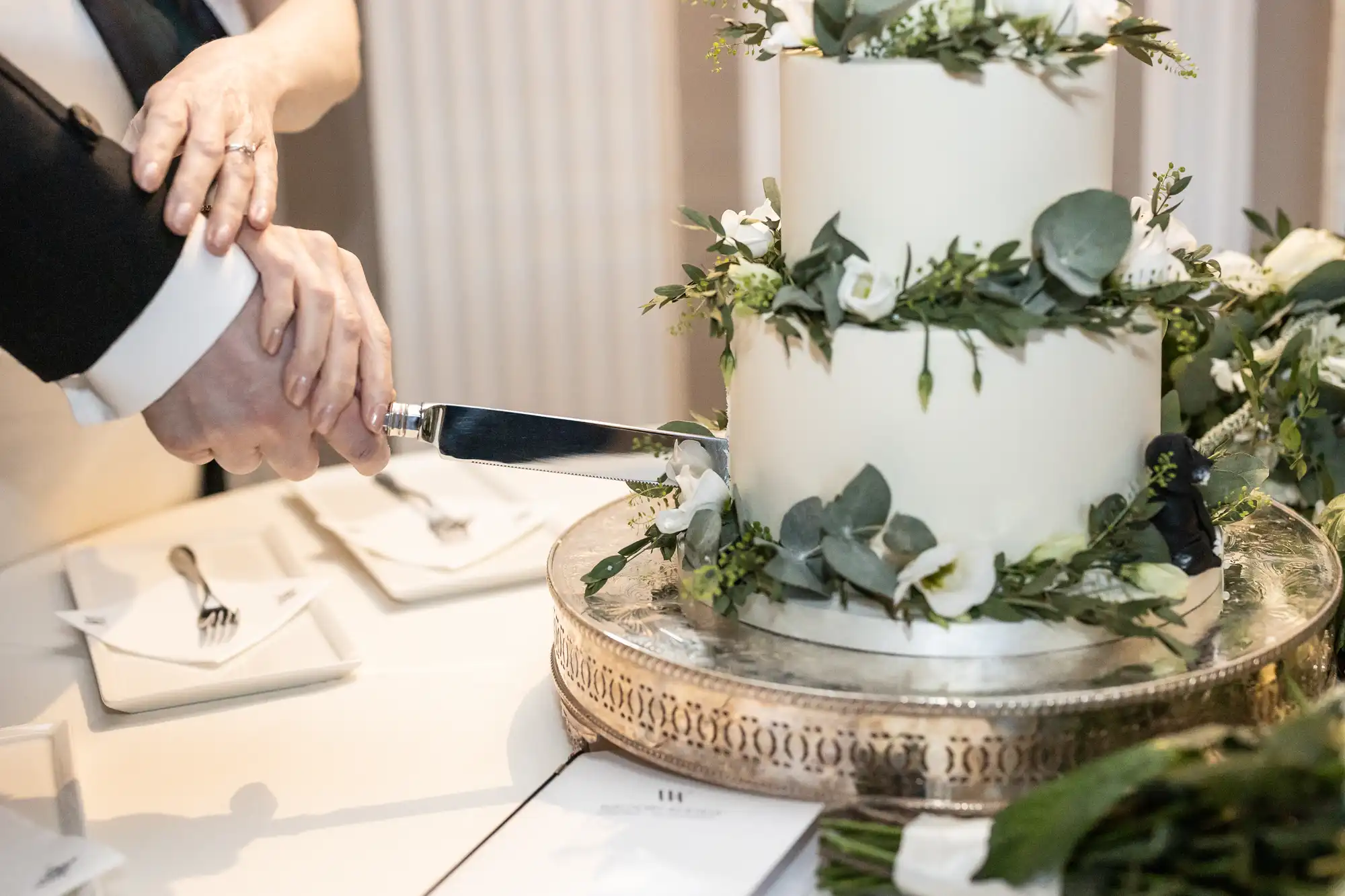 A couple cuts into a white, two-tiered wedding cake decorated with greenery and flowers. The cake sits on an ornate silver stand.