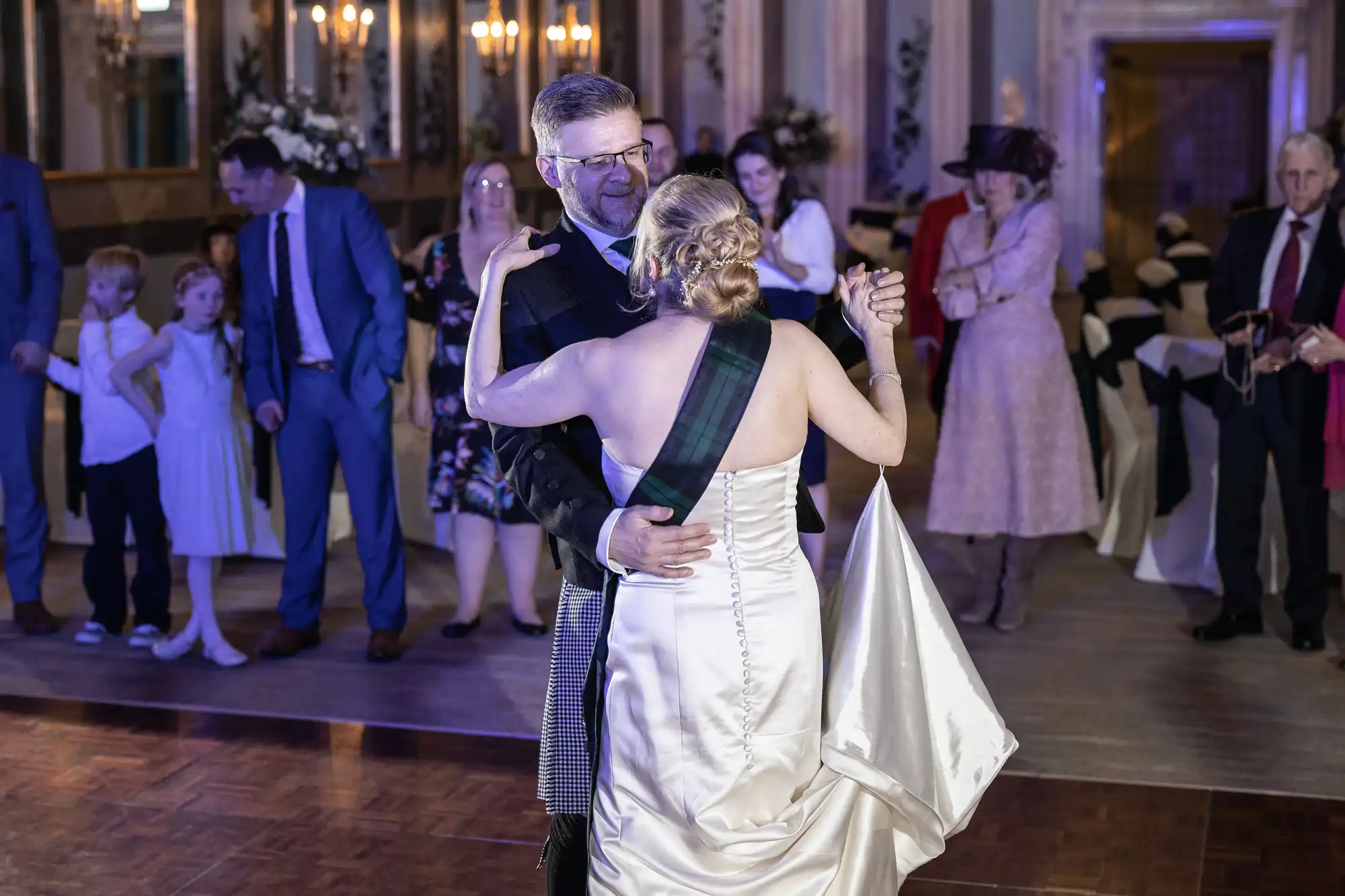 A couple dances closely in formal attire at an indoor event while onlookers in the background watch and converse.