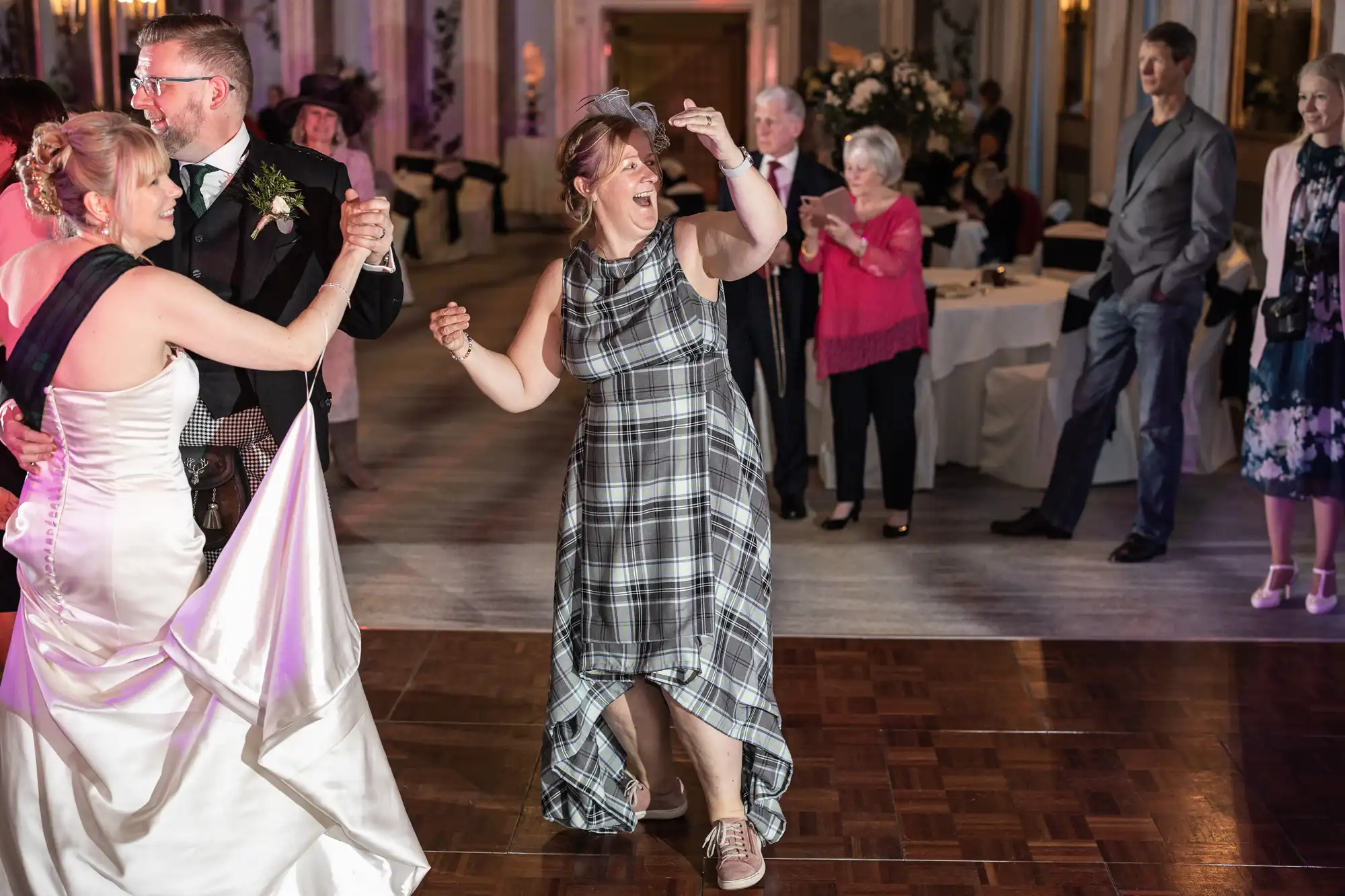 A bride and groom dance at their wedding reception while a guest in a plaid dress and sneakers energetically joins them. Additional guests observe from nearby.