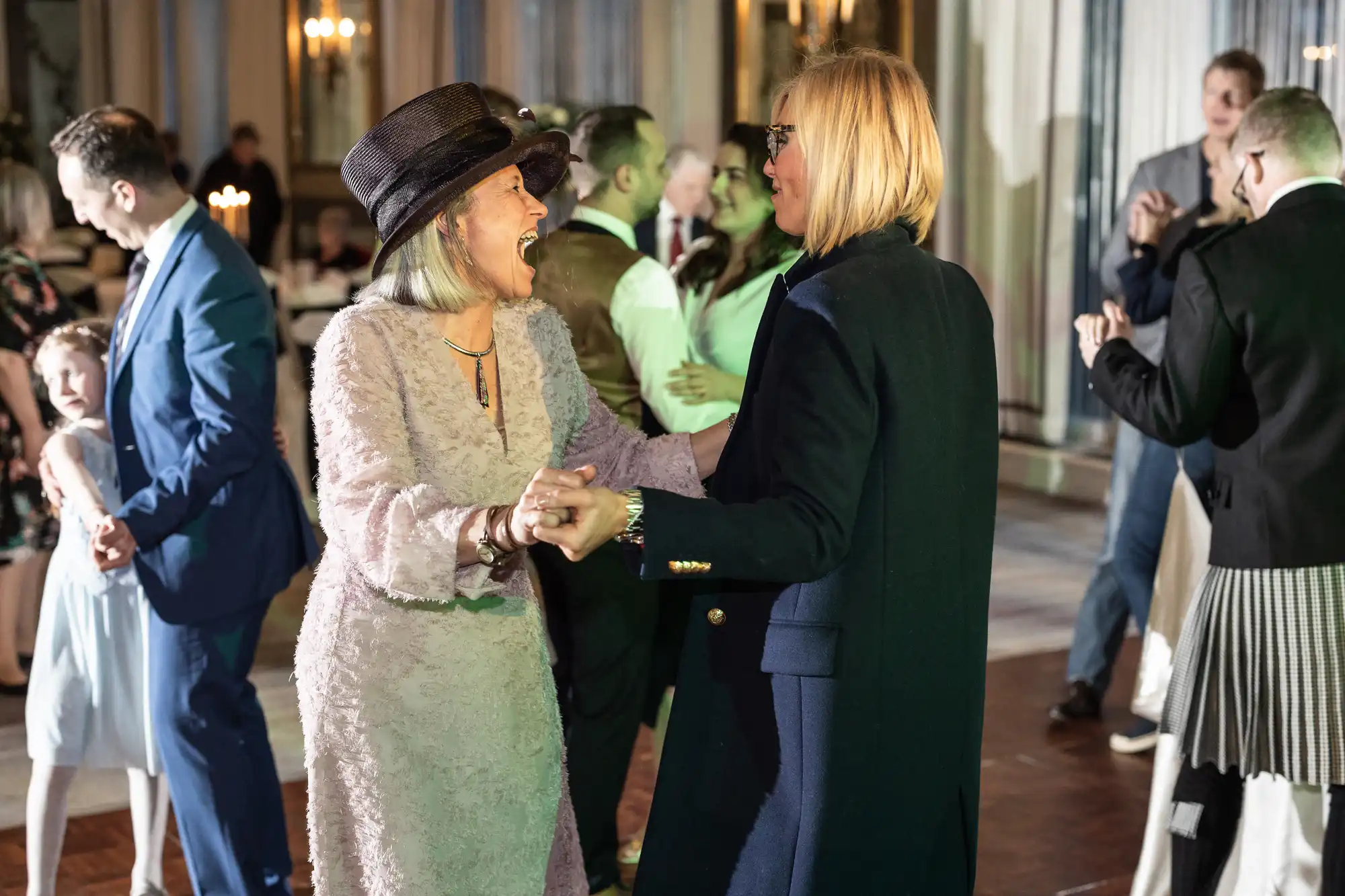 Two women joyfully dance together at a formal event. One wears a pink lace dress and a wide-brimmed hat; the other dons a dark coat. People in the background are also dancing and mingling.