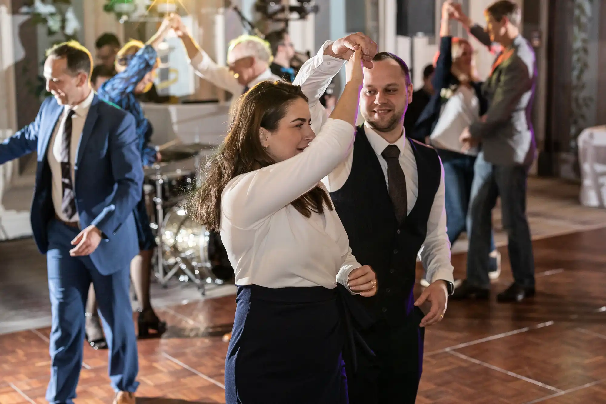 A group of people in formal attire are dancing at an indoor event. A man in a vest is twirling a woman in a white blouse on the dance floor. A drum set is visible in the background.