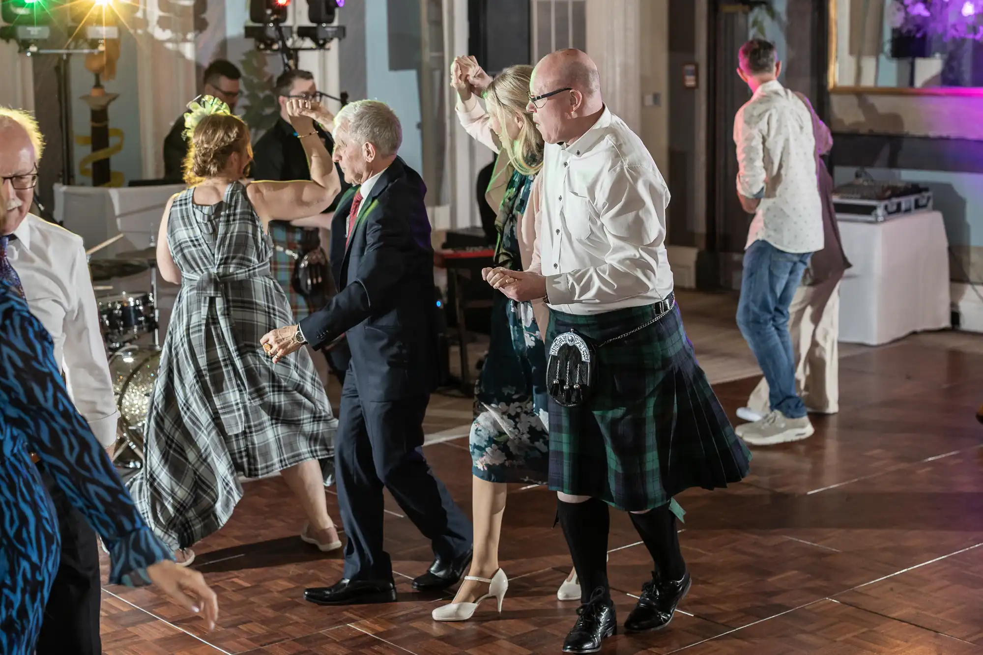 A group of people dressed in formal attire, including kilts, dancing energetically on a wooden dance floor at an indoor event with musical equipment visible in the background.