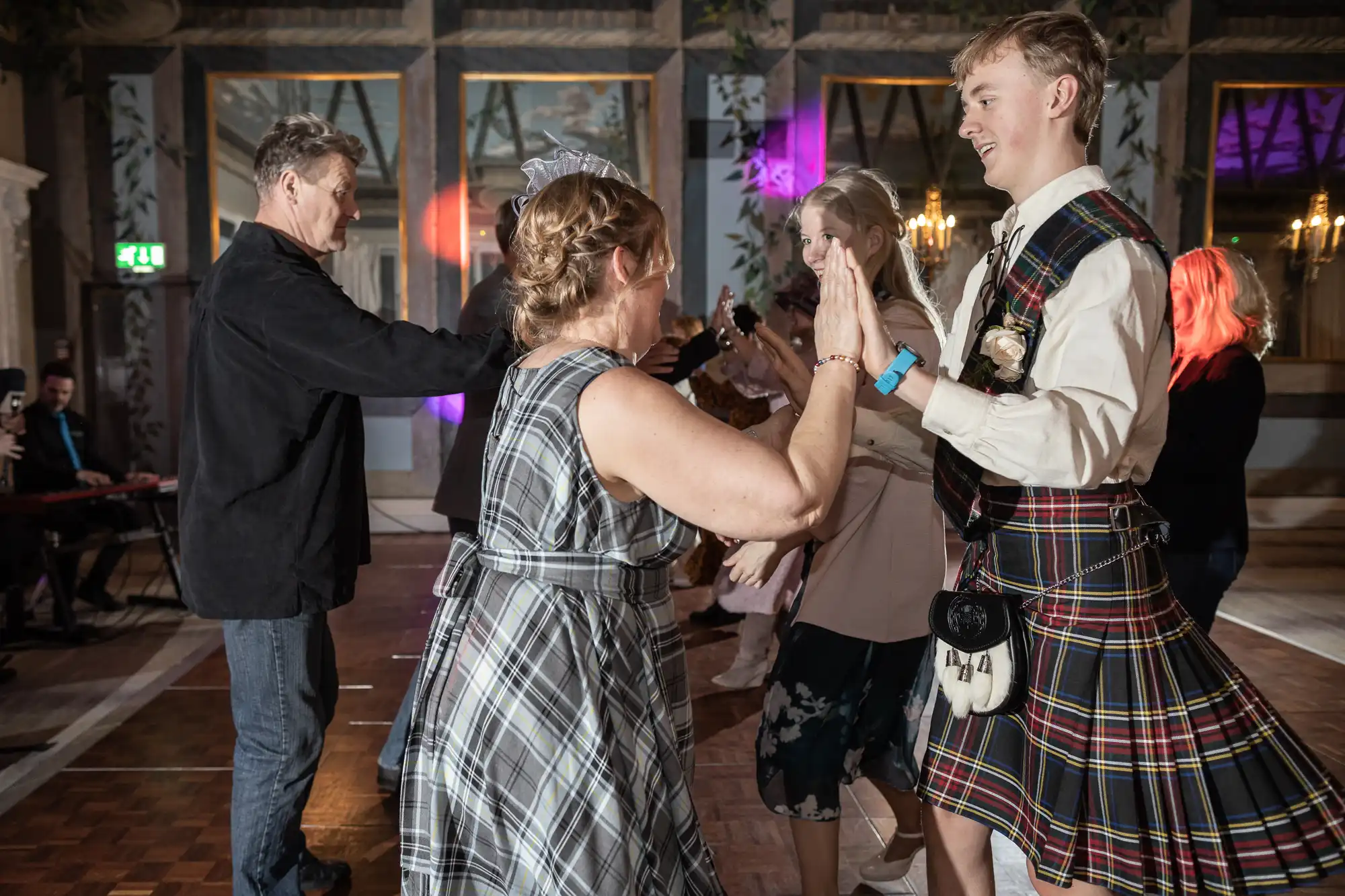 People dressed in formal attire are dancing in pairs at an indoor event. A man in a kilt and a woman in a plaid dress are facing each other, clapping hands. Other pairs dance in the background.