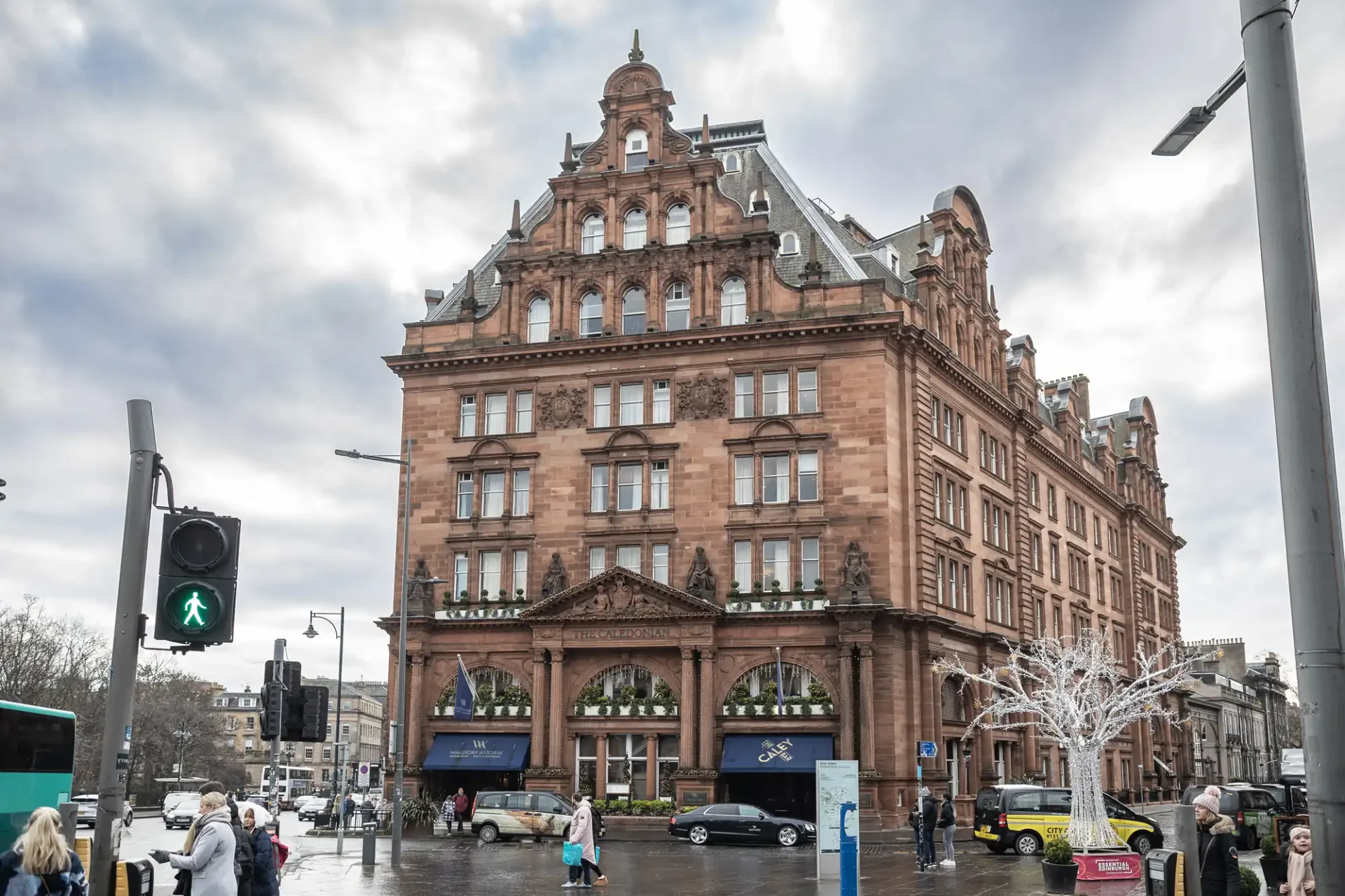 A large, historic, red-brick building on a busy city street corner, with pedestrians, a traffic light, and a few parked cars visible, under a cloudy sky.