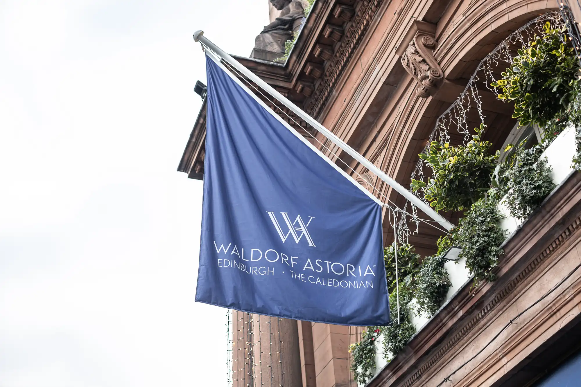A blue flag with white text reading "Waldorf Astoria Edinburgh - The Caledonian" is mounted on the exterior of a historic building adorned with greenery.