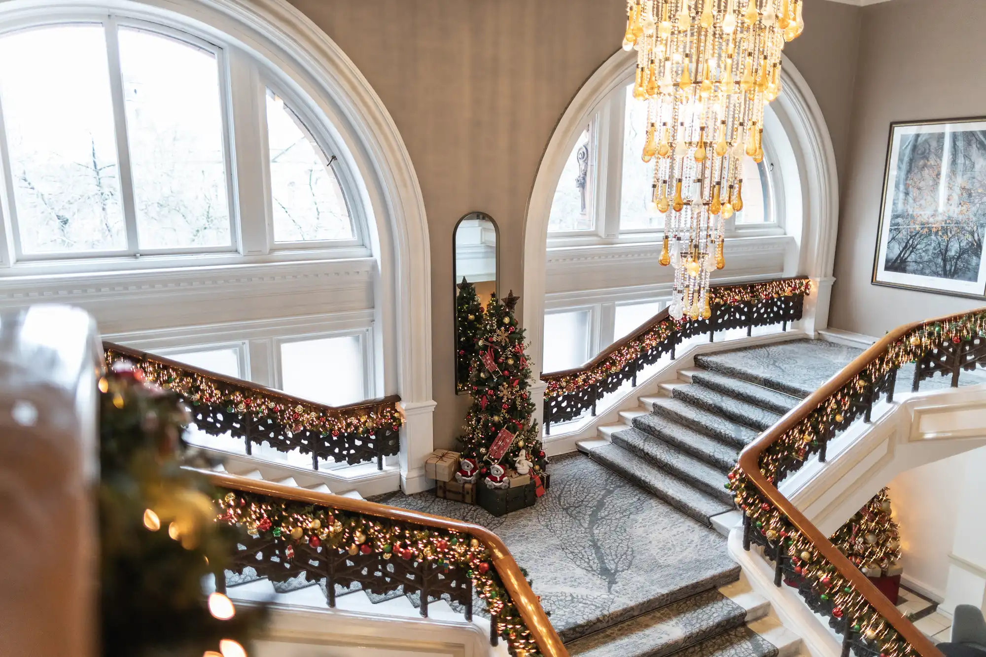 A grand staircase decorated with garlands and lights, featuring a large central window, chandelier, and a Christmas tree at the base, inside an elegant interior space.