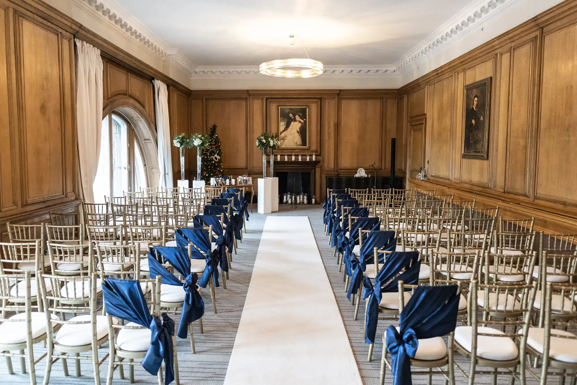 A neatly arranged wedding ceremony setup in an elegantly wooden-paneled room. Rows of gold chairs, some with blue bows, line a white aisle runner leading to an altar with flower arrangements.