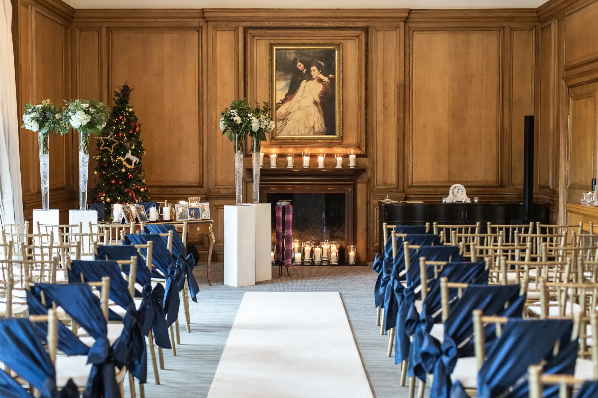 A wedding ceremony setup with rows of chairs adorned with blue bows facing an altar. The altar features floral arrangements, candles, and a painting above a fireplace. A decorated Christmas tree is visible.