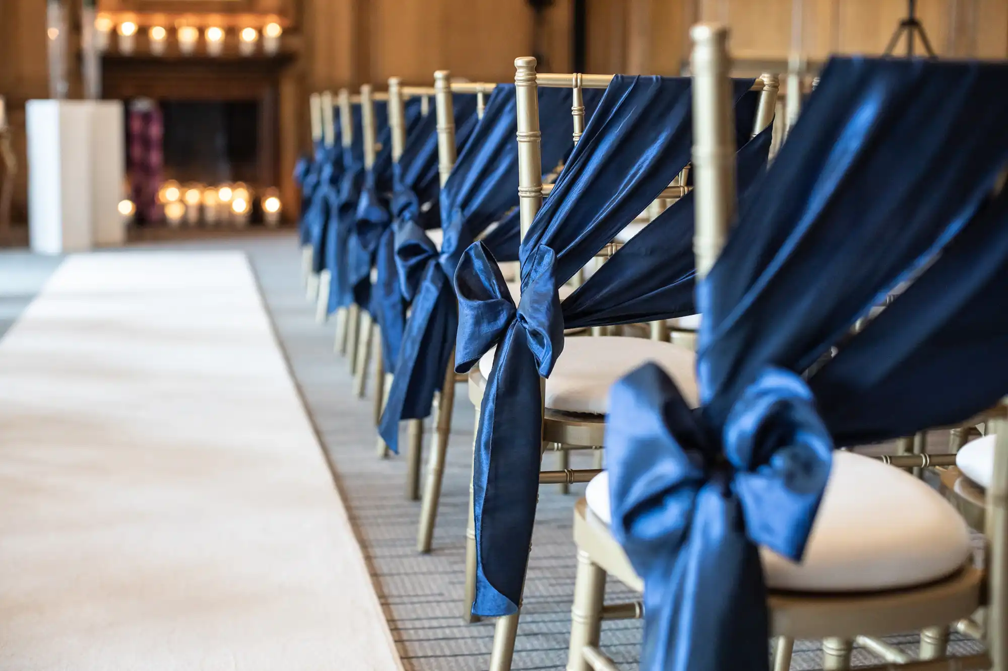 Rows of chairs with blue bows are aligned next to a white aisle, set up for an indoor event. Candles are lit on a mantel in the background.