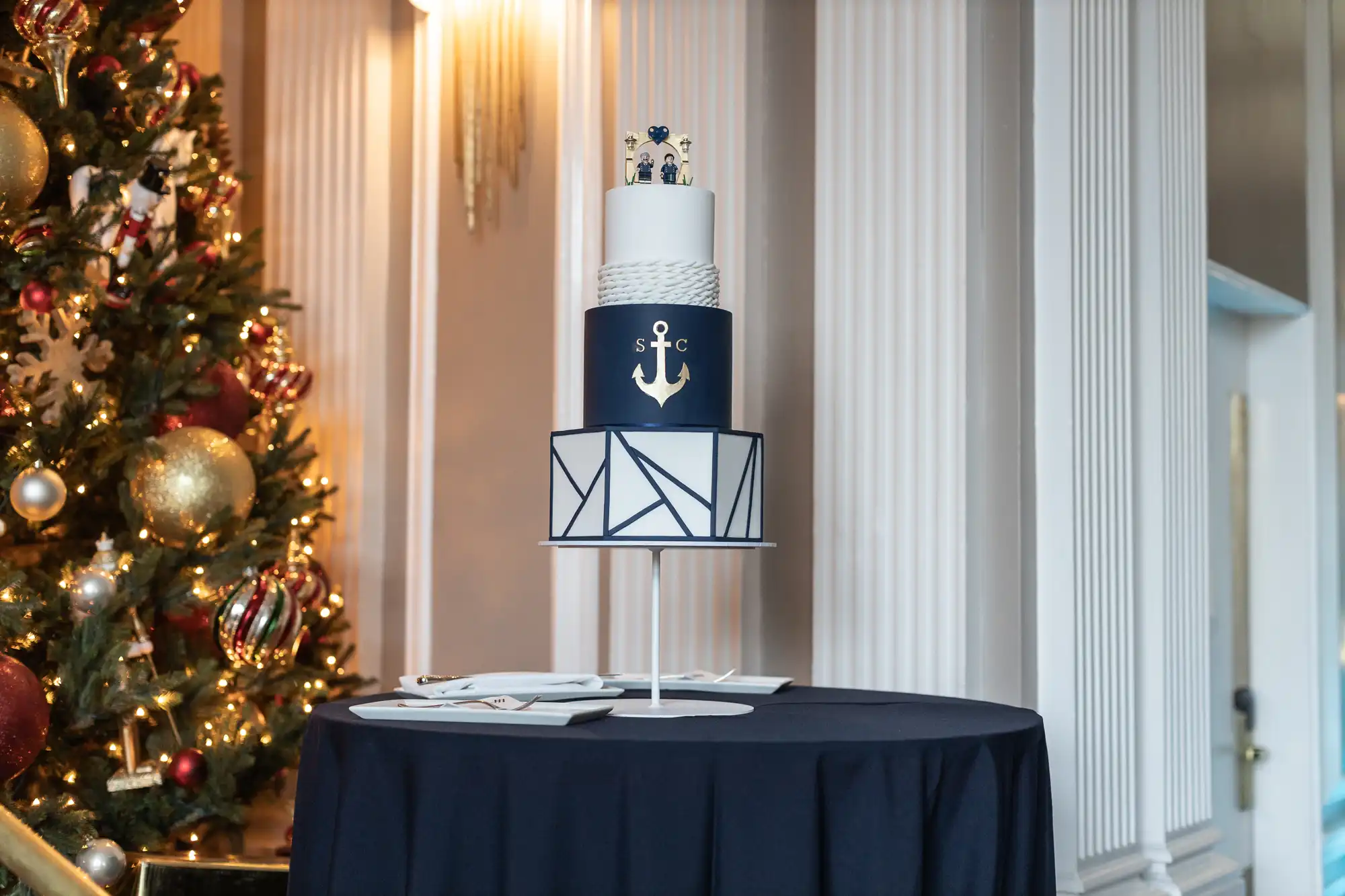 A three-tier navy and white cake with an anchor design sits on a round table, which is covered with a dark tablecloth. A decorated Christmas tree is seen in the background on the left.