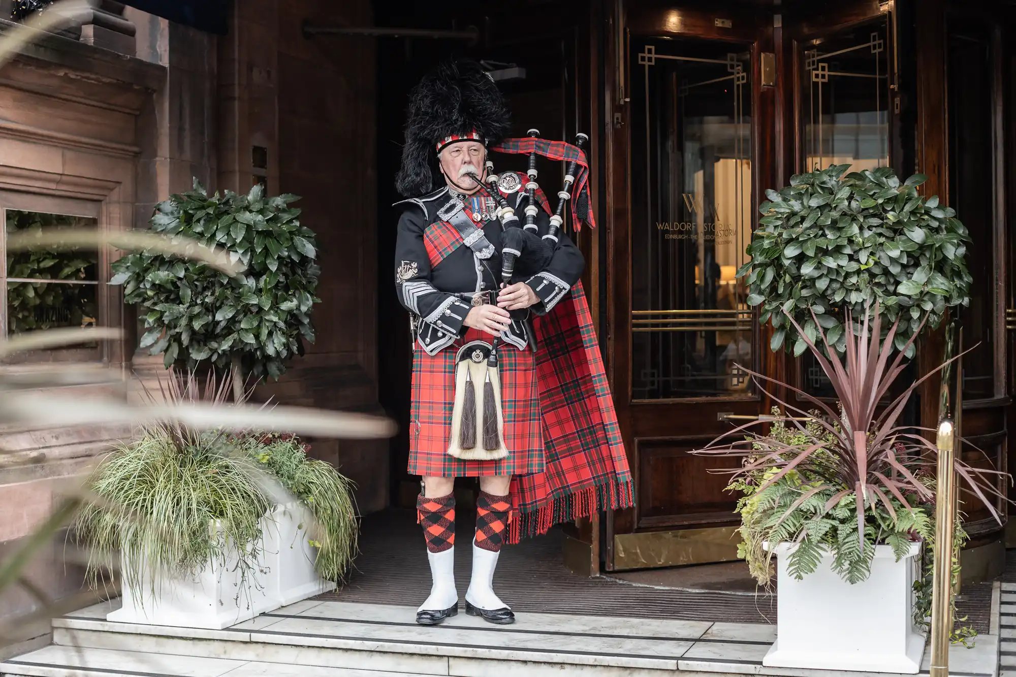 A man in traditional Scottish attire plays the bagpipes outside a building entrance, flanked by two potted plants.