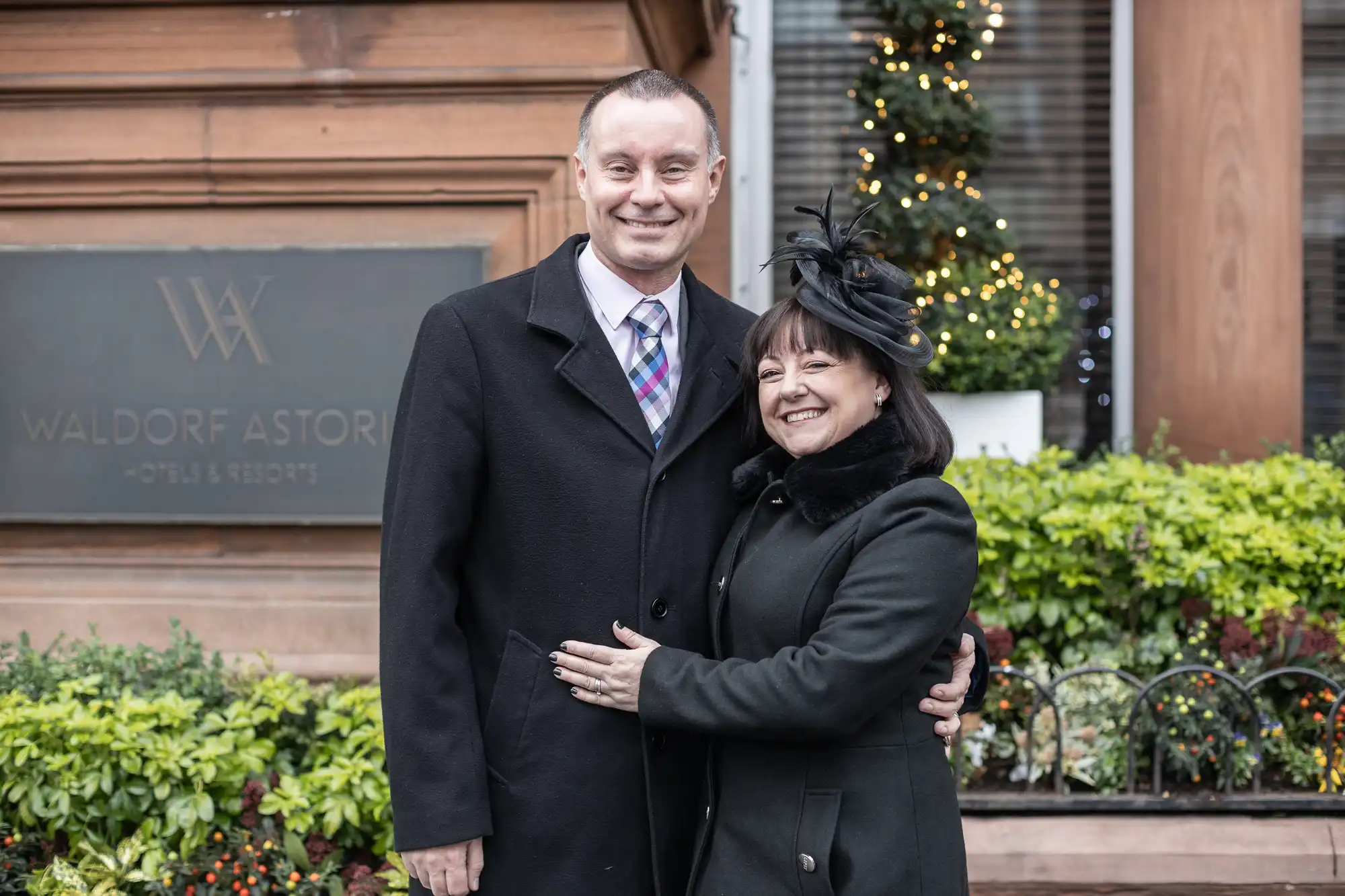 A man and a woman, both dressed in coats, pose and smile together in front of a Waldorf Astoria Hotels & Resorts sign, with greenery and a festive decoration in the background.
