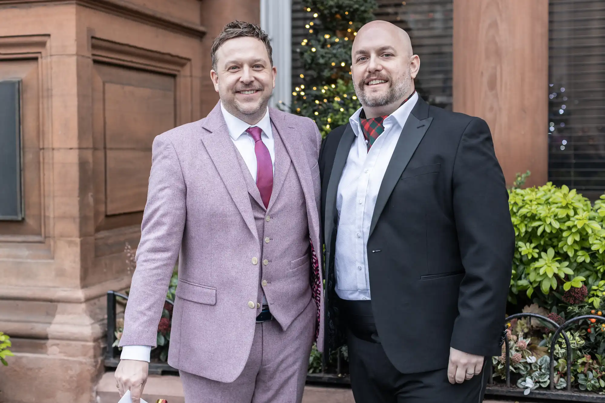 Two men standing side by side, one wearing a light pink suit with a red tie and the other in a dark suit with a patterned tie, in front of a building with plants and a decorated tree in the background.