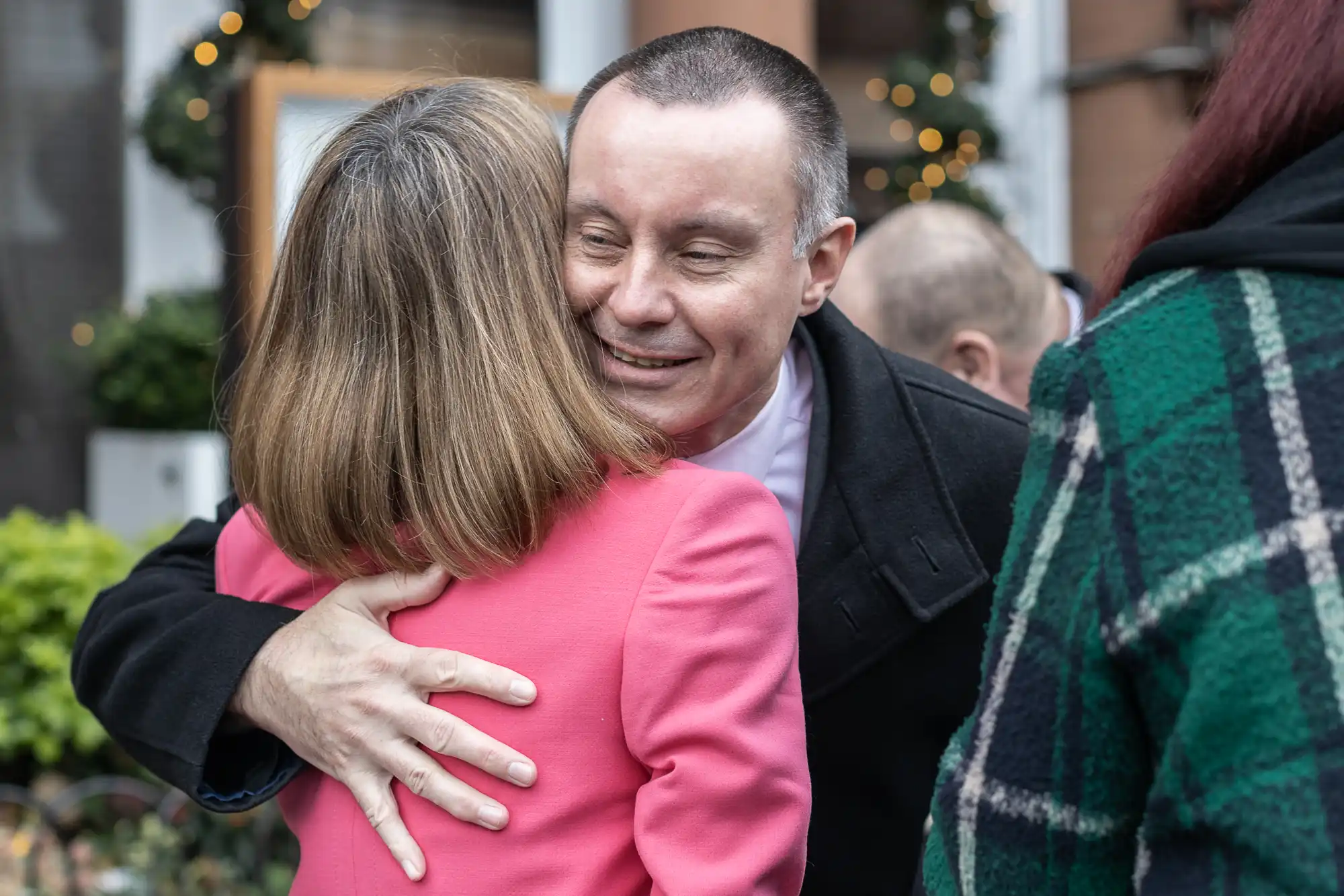 Two people are hugging outside in a festive setting. The person facing the camera is smiling warmly, while the other has their back turned, wearing a pink jacket. Other people are visible in the background.