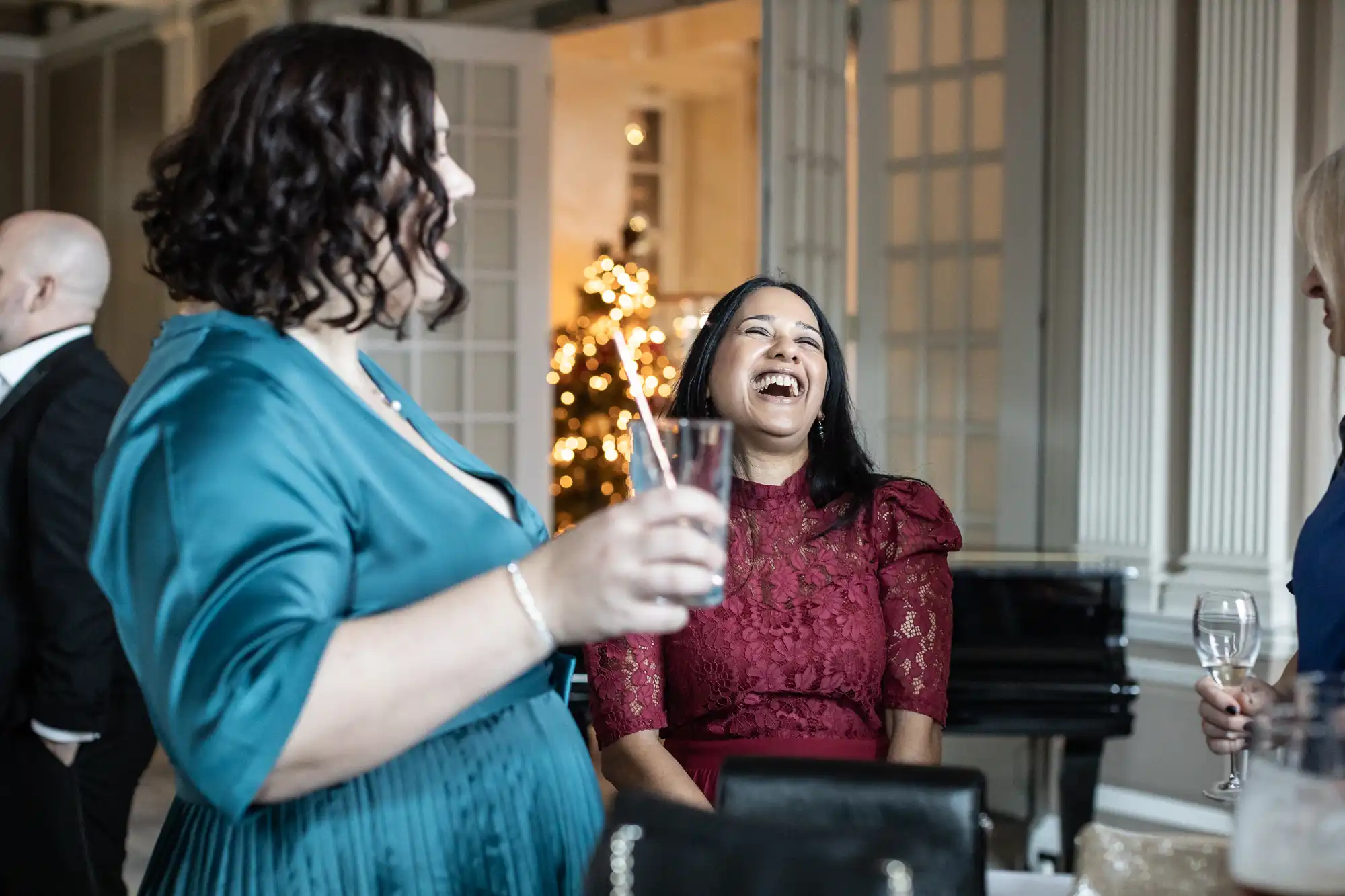 Two women are enjoying drinks and laughing together indoors, decorated with a Christmas tree in the background.