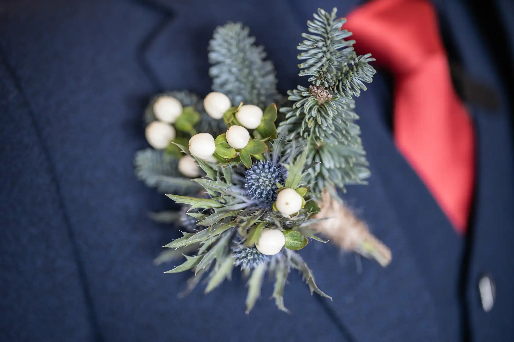 Close-up of a man's suit jacket with a floral boutonniere made of greenery, white berries, and small blue flowers pinned to the left side lapel. He wears a red tie.