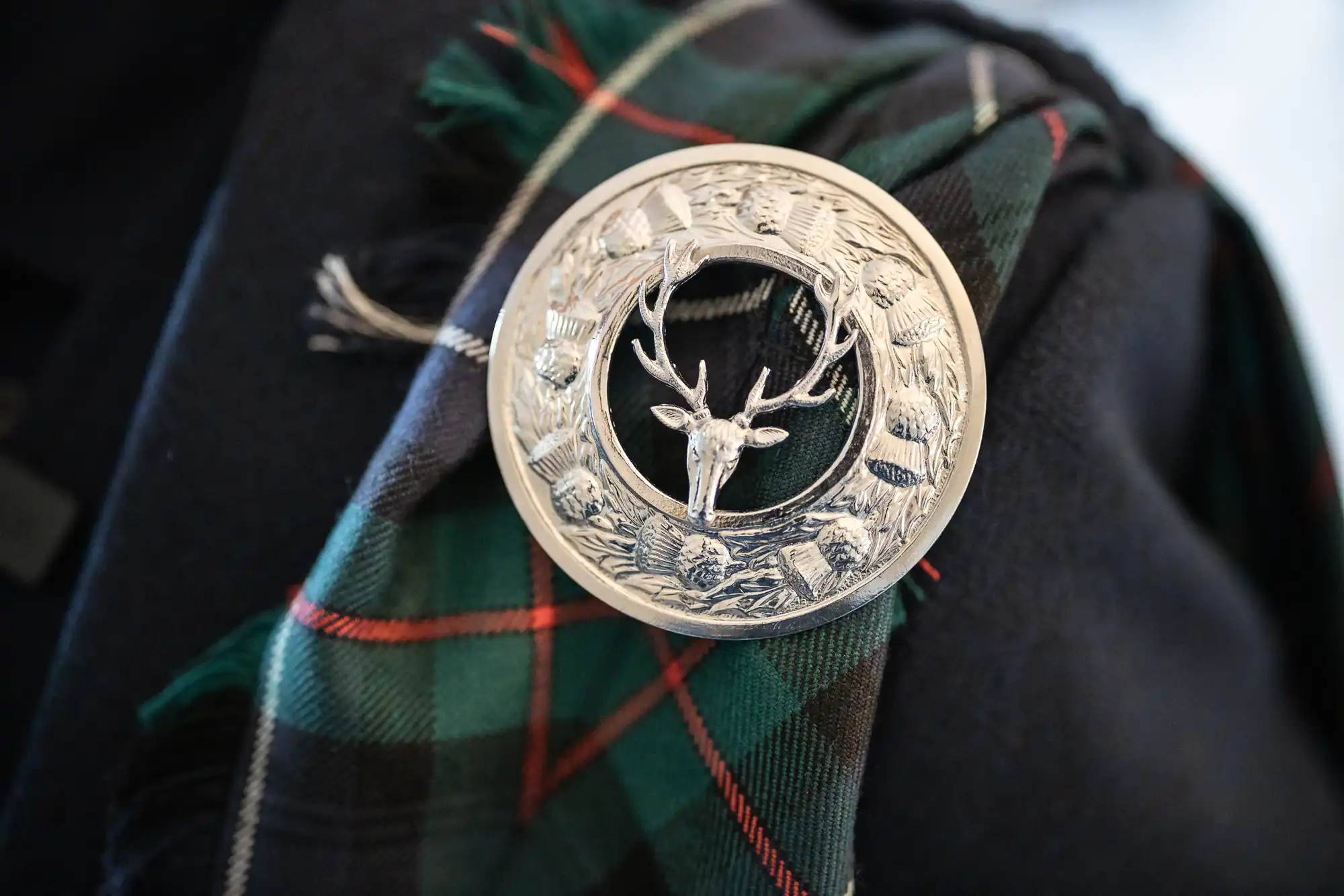 A silver brooch featuring the head of a deer is pinned to a tartan fabric, showcasing a traditional Scottish design.