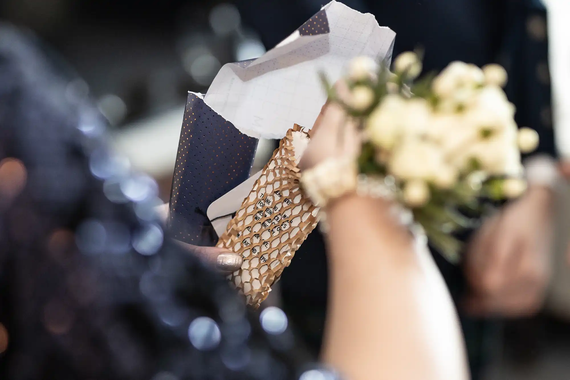 A person opens a small, intricately wrapped gift while holding a bouquet of white flowers.