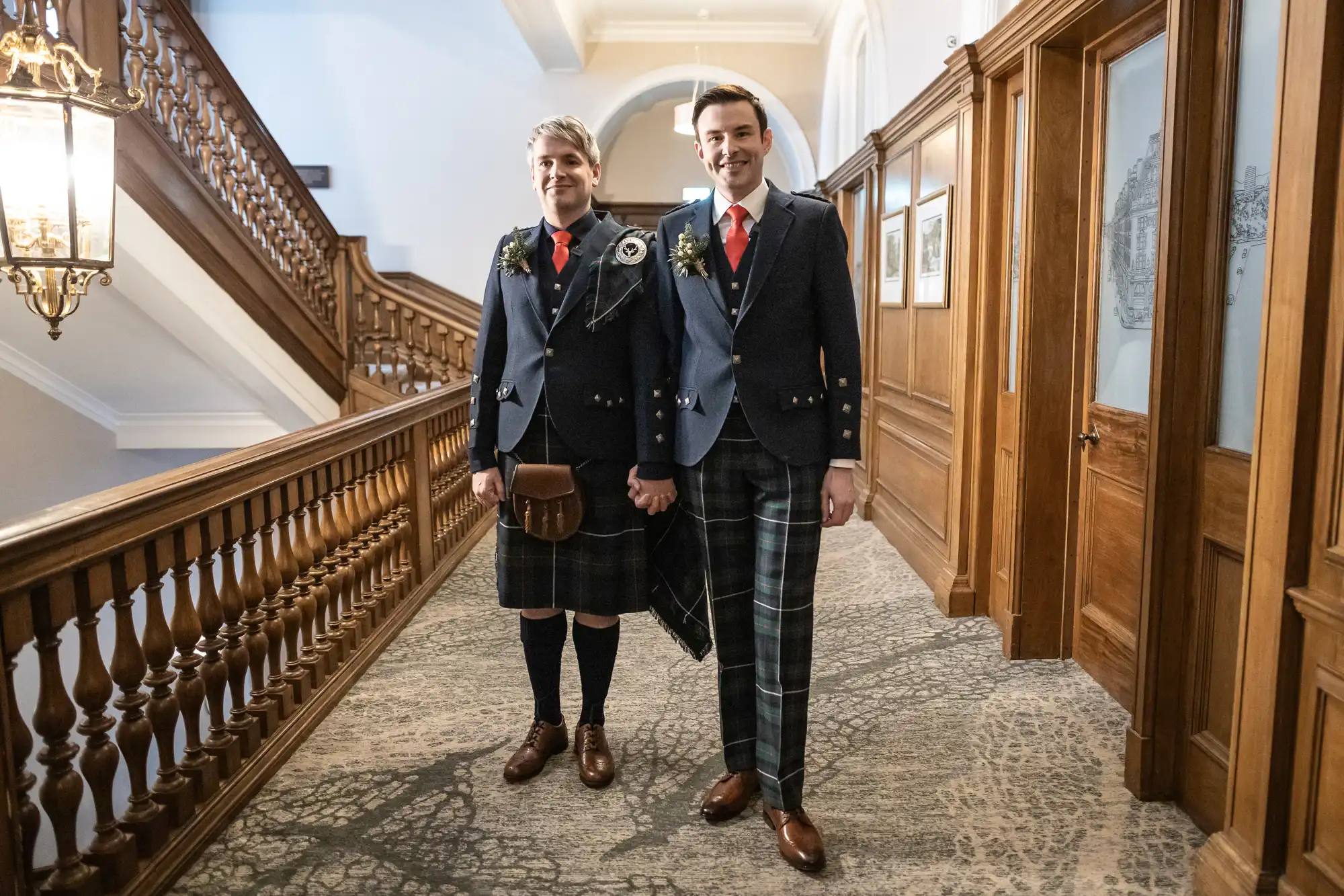 Two men stand in a hallway wearing traditional Scottish kilts and jackets, smiling at the camera.