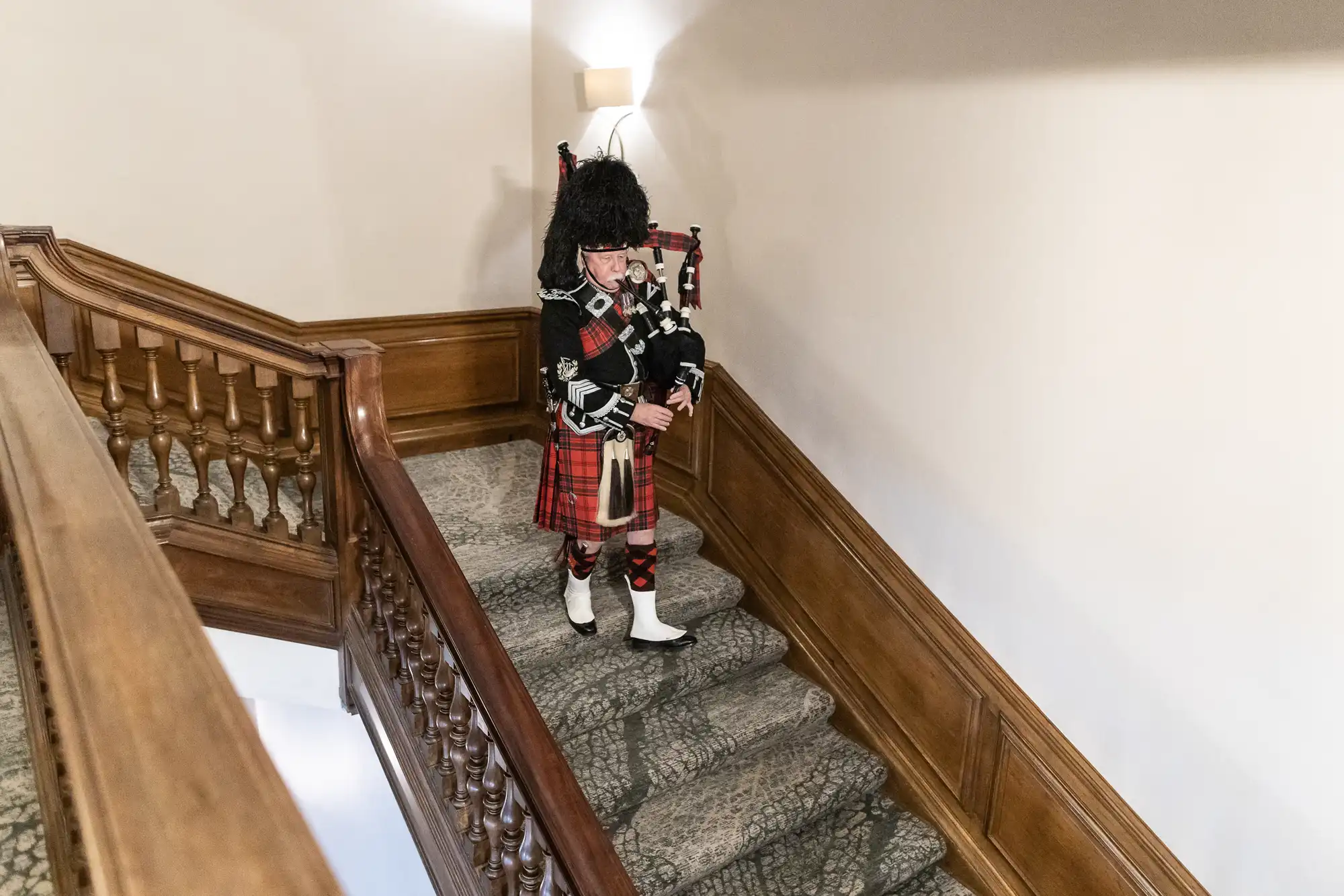 A person dressed in traditional Scottish attire, including a kilt and a feathered hat, plays bagpipes while descending a wooden staircase.
