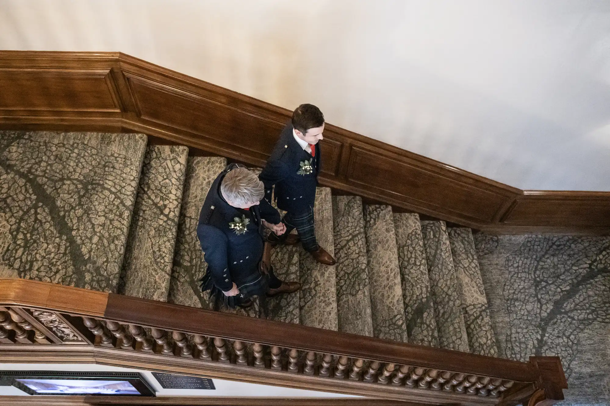 Two men in formal attire descend a carpeted staircase with wooden bannisters.
