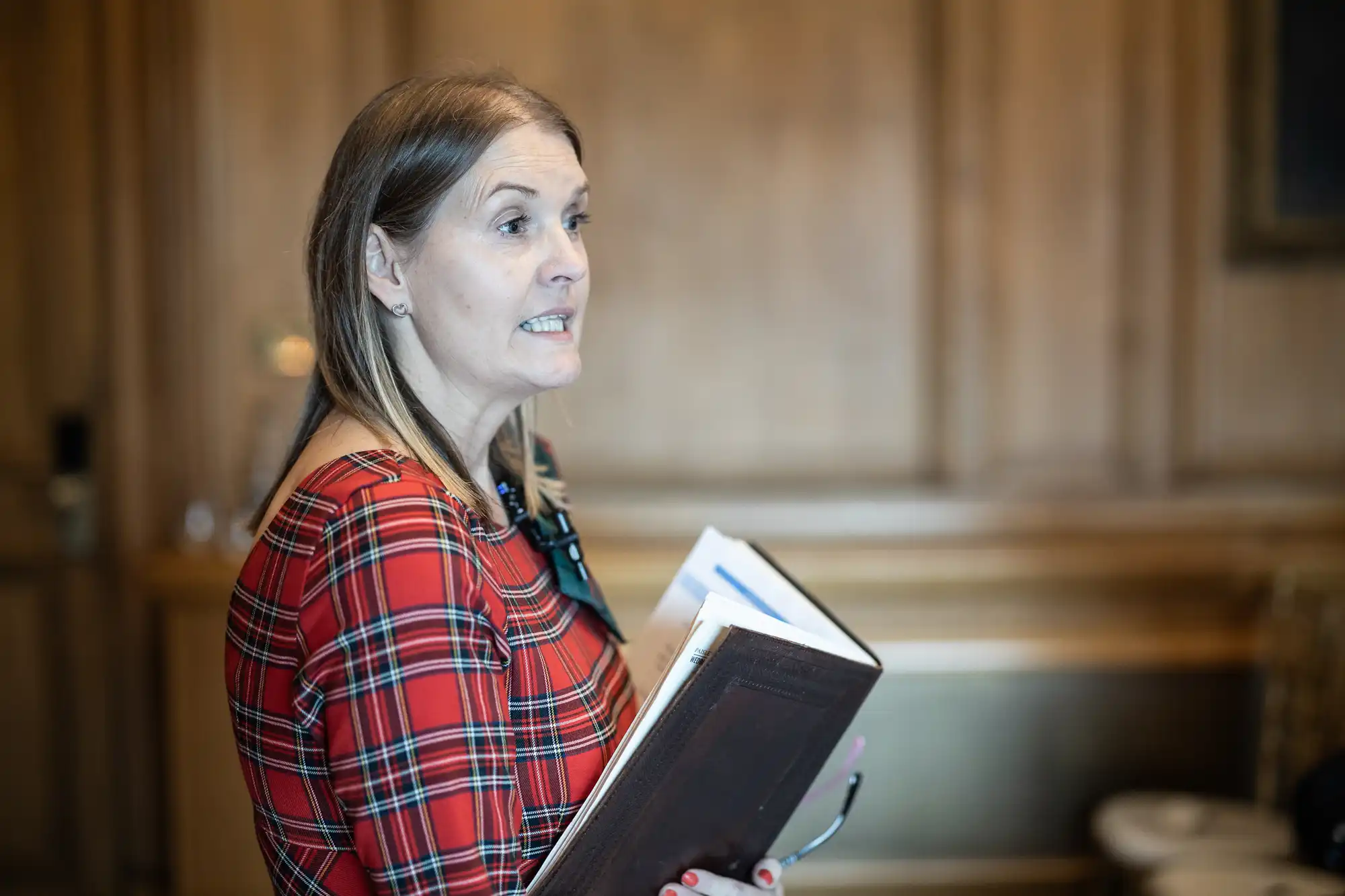 A person with shoulder-length hair wearing a red plaid outfit is holding a few books and papers while speaking in a wood-paneled room.