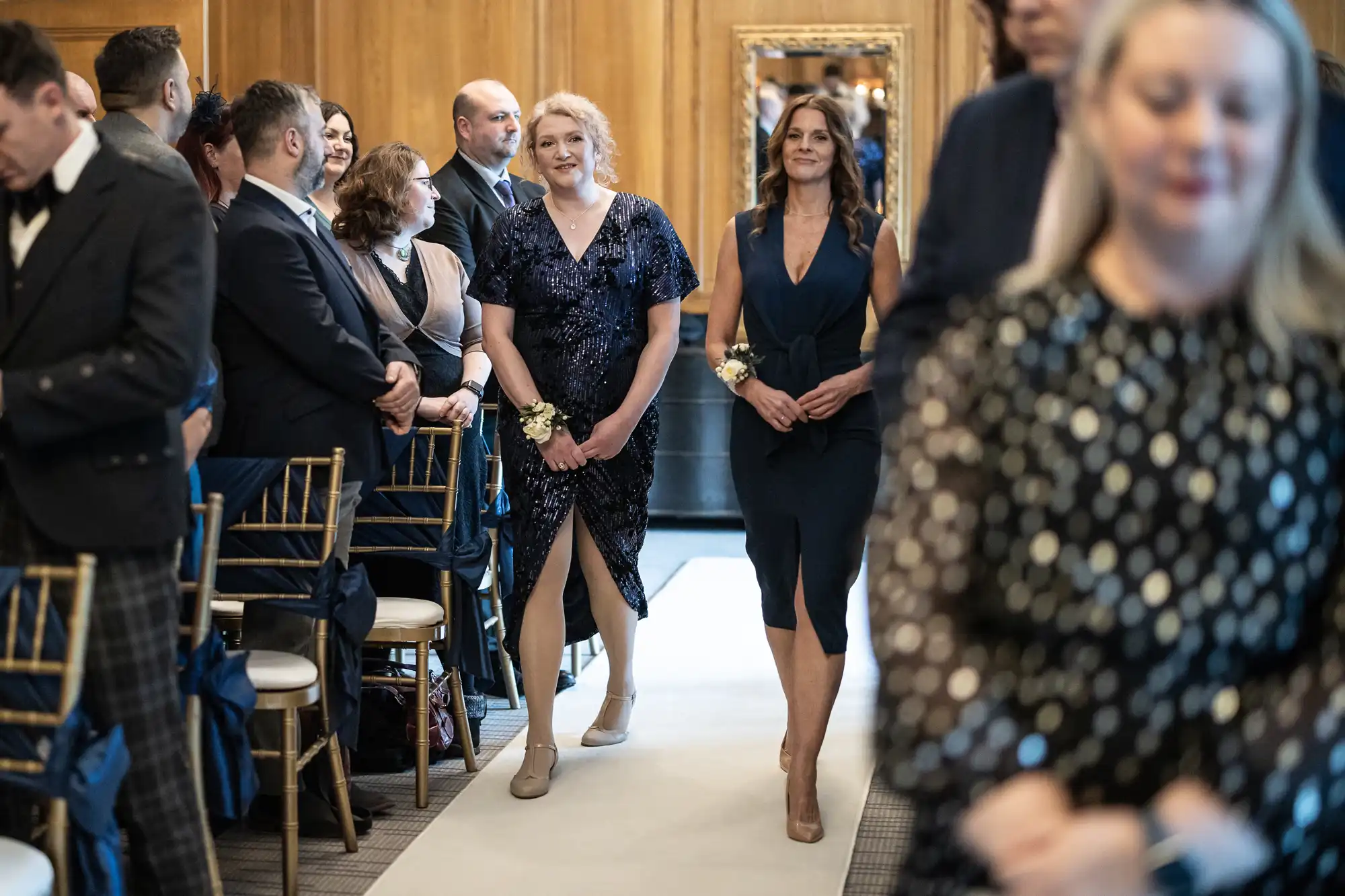 Two women walk down an aisle during a formal event, while guests seated on either side watch. The surroundings feature wooden paneling and elegant decor.