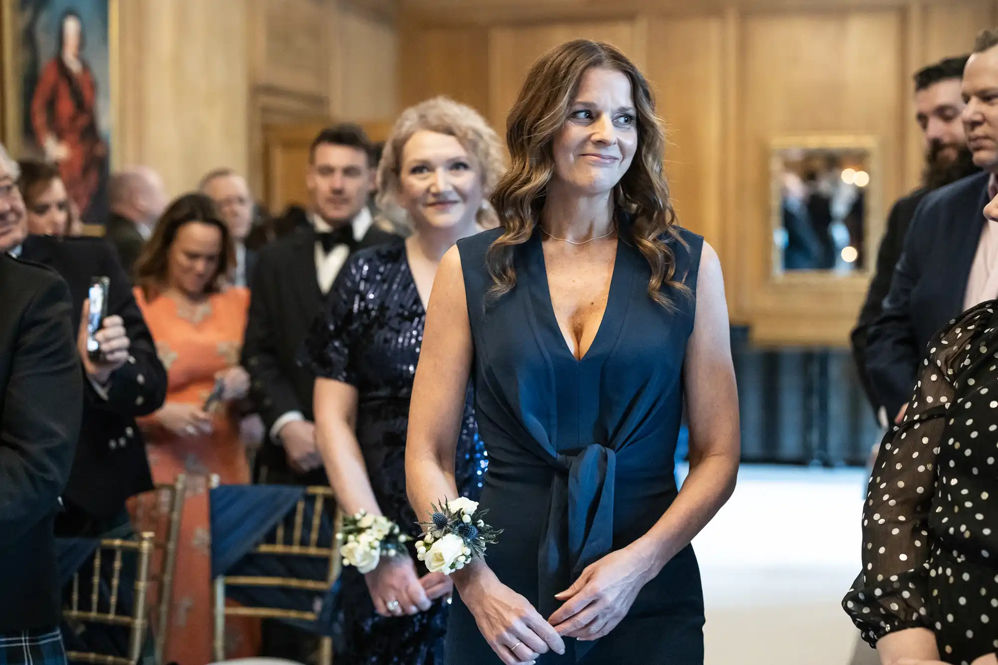 A woman in a navy dress with a flower corsage walks down an aisle with seated guests smiling and looking on in a formal indoor setting.