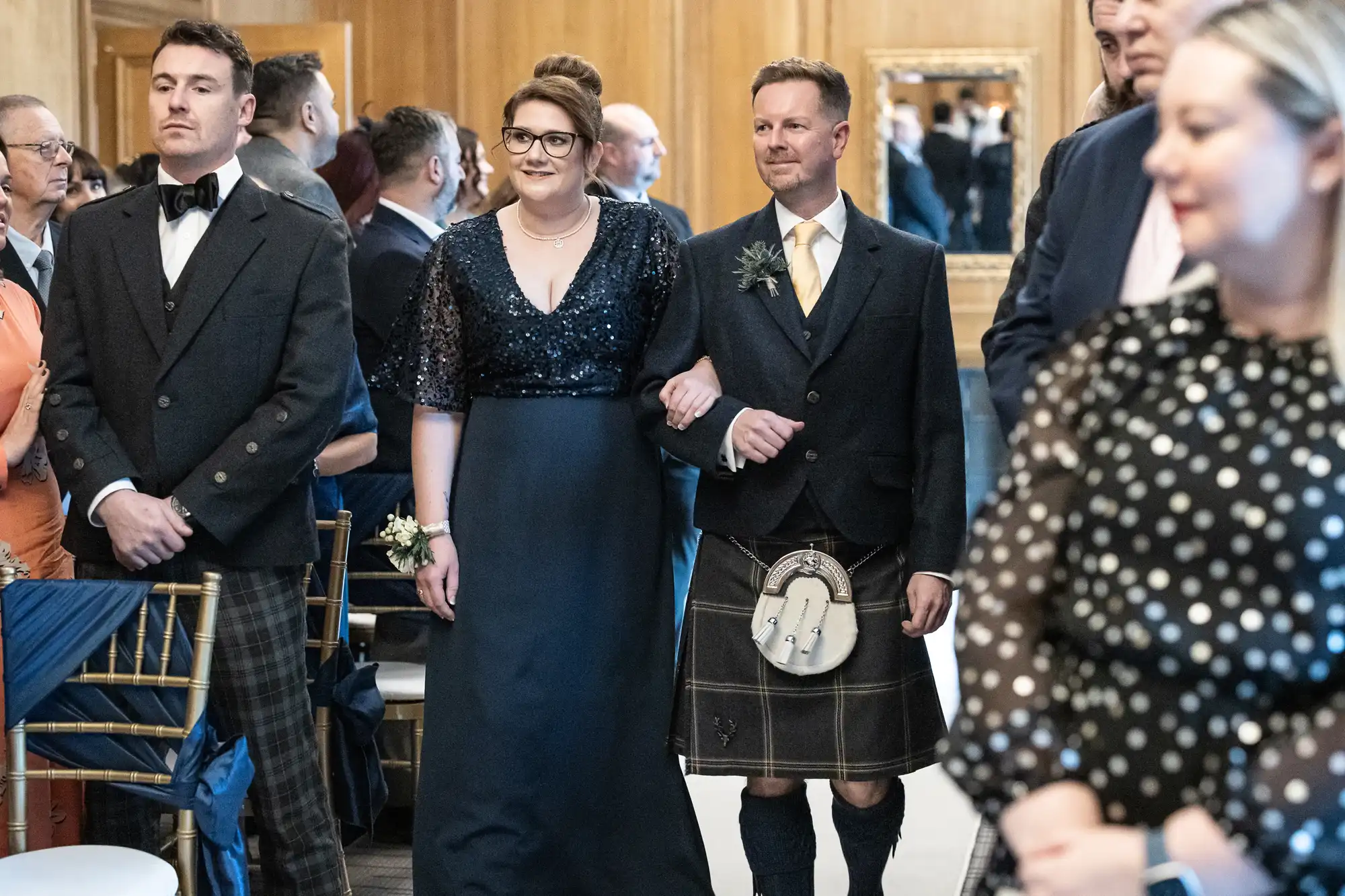 A man and woman, formally dressed, walk arm in arm down an aisle. The man wears a traditional Scottish kilt and sporran, while the woman wears a dark dress with sparkly details. Others look on in the background.