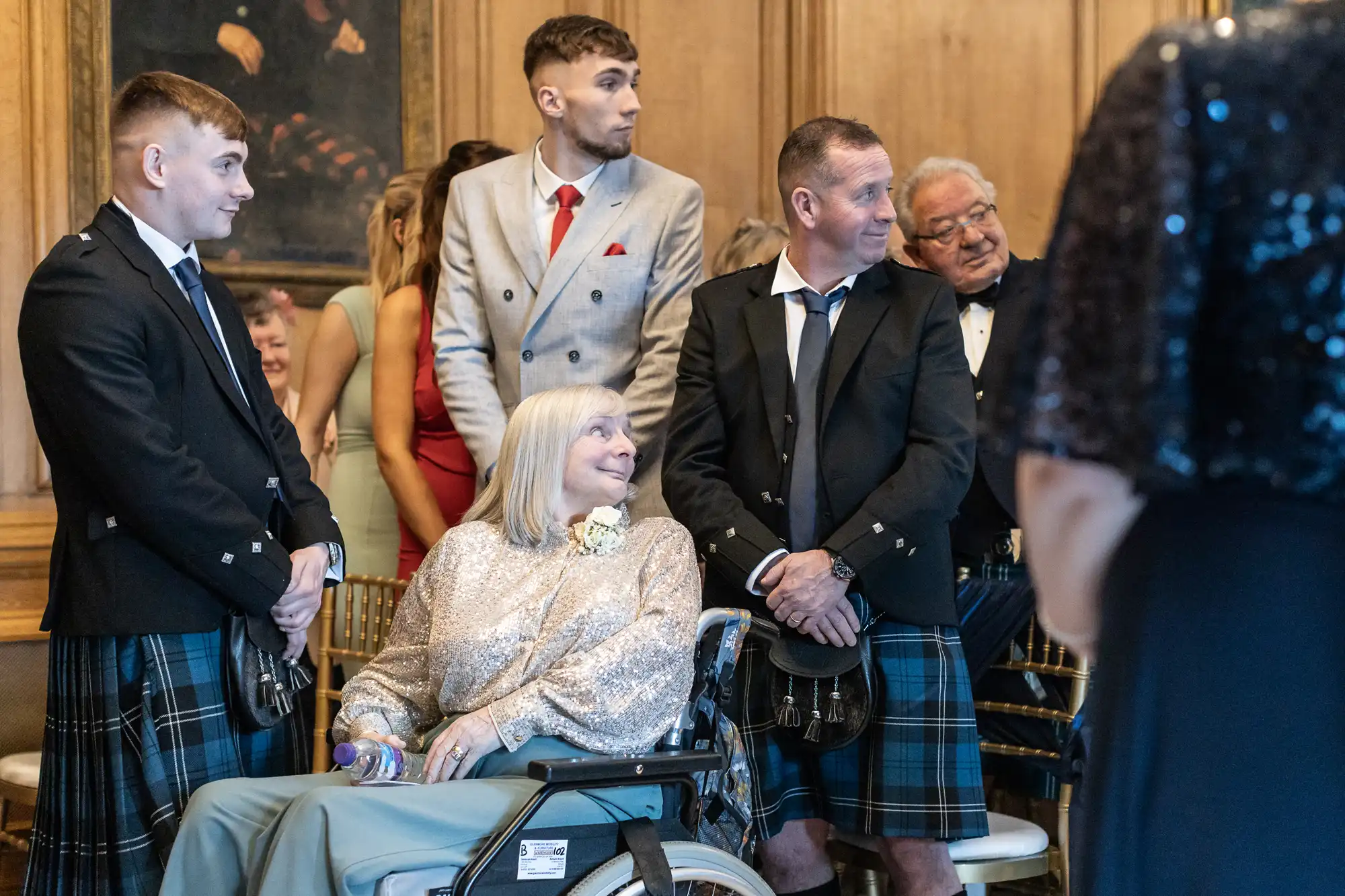 A group of people in a formal setting. A woman in a wheelchair is in the center, surrounded by men wearing kilts and jackets. They are engaged in conversation, with others in the background.