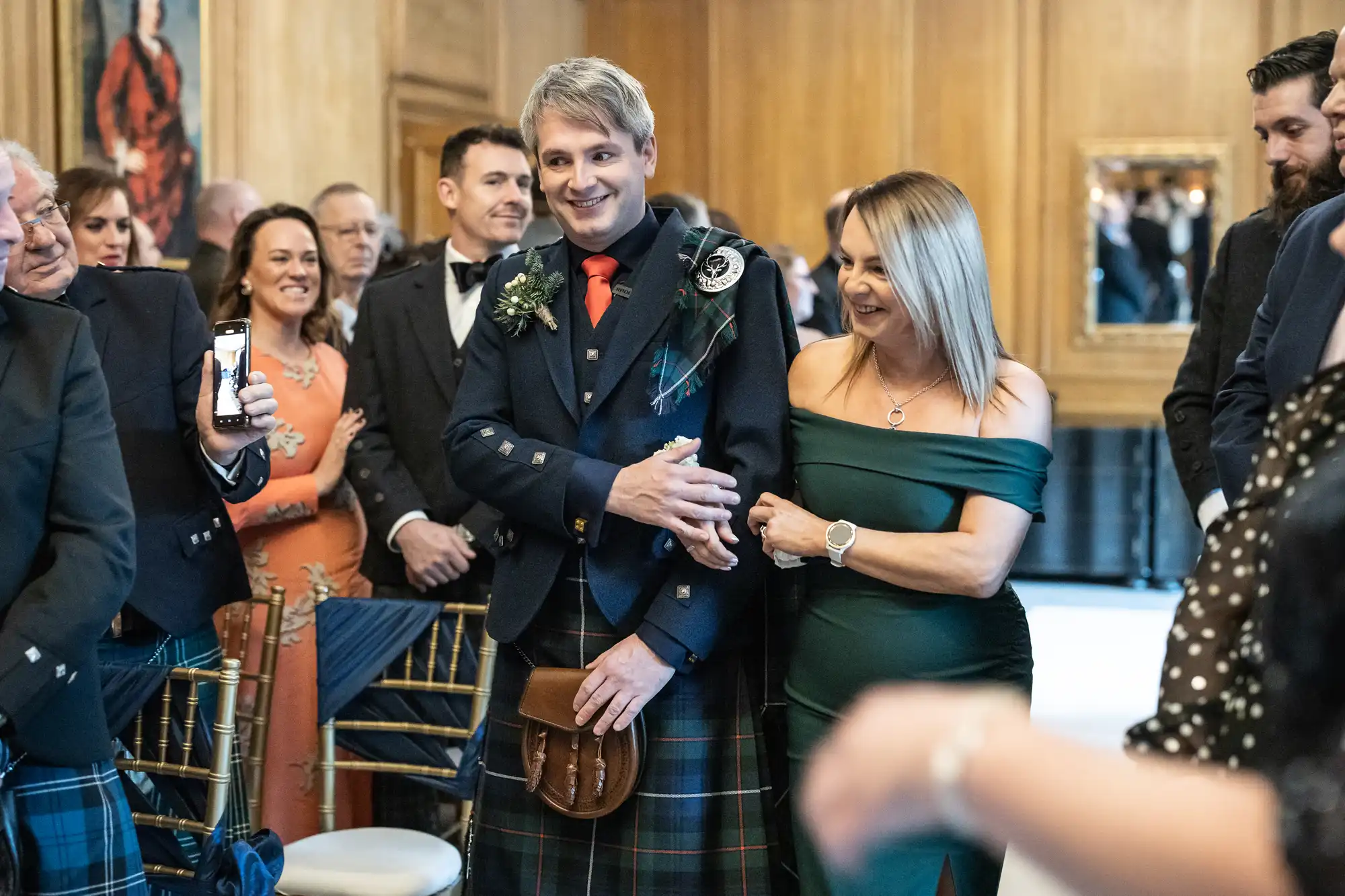 A person in Scottish attire walks down an aisle arm-in-arm with another individual in a green dress, surrounded by smiling onlookers in a formal setting.