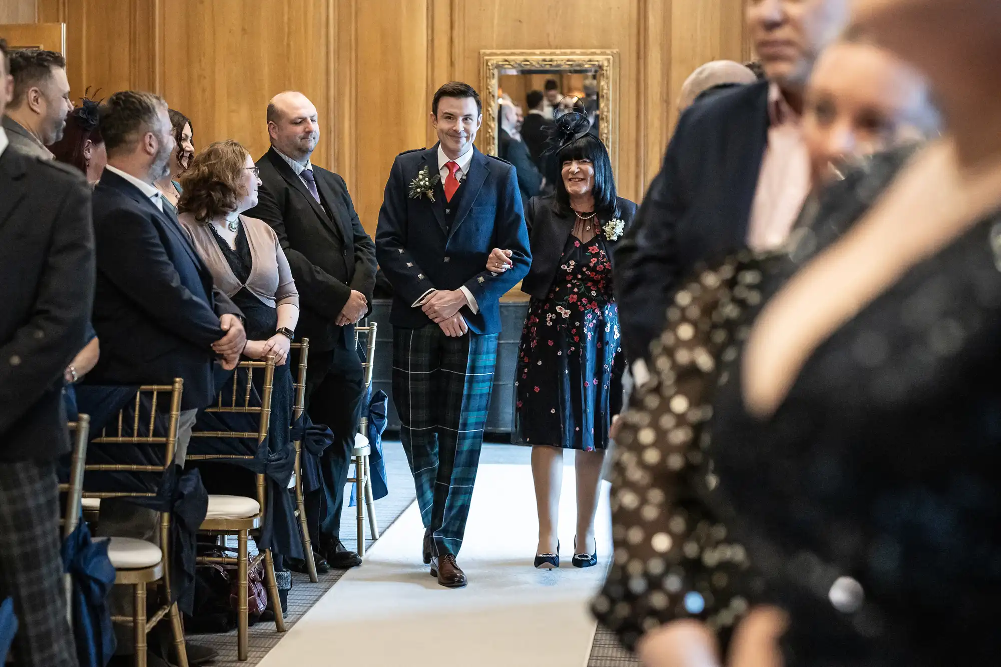 A man in a suit and plaid pants walks down an aisle with an older woman in a gown. They are surrounded by seated guests in a wooden paneled room.