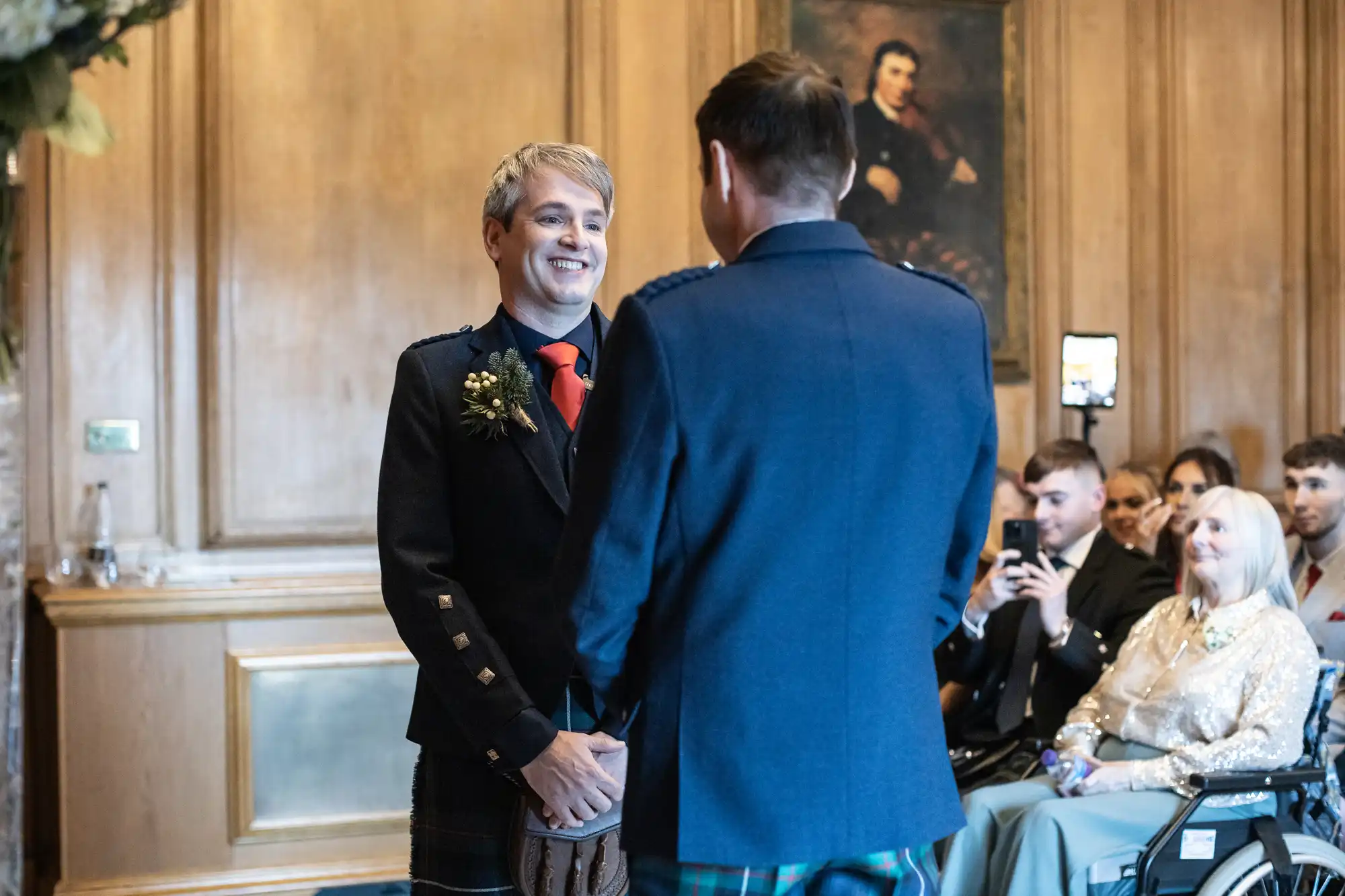 Two men in traditional Scottish attire stand facing each other during a wedding ceremony. Guests are seated in the background, and a person captures the moment on a smartphone.