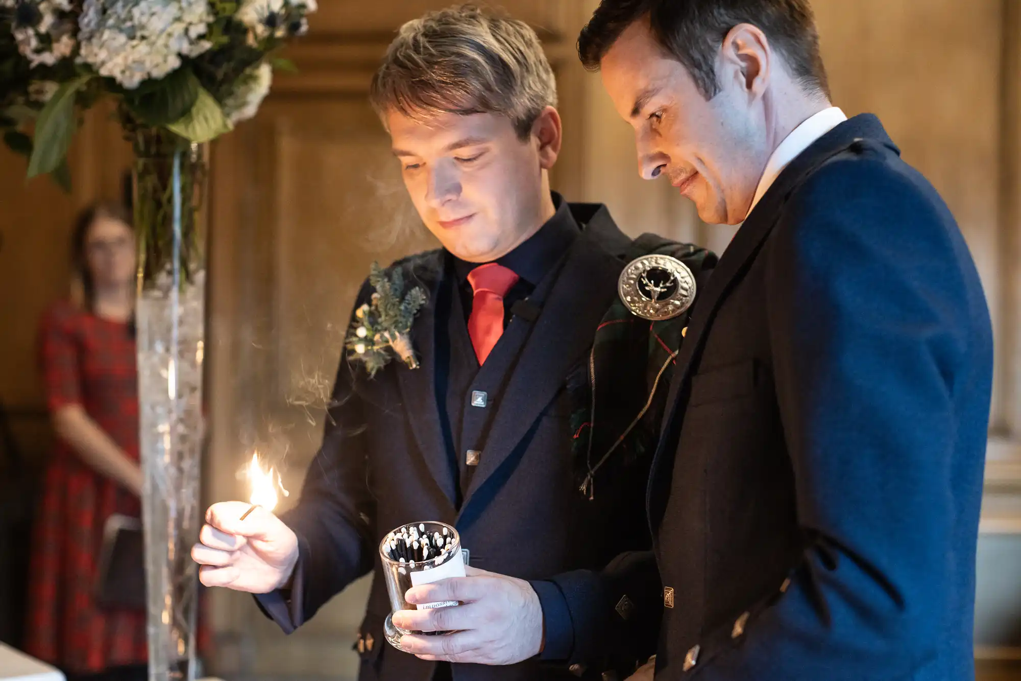 Two men stand together, one lighting a candle while the other watches closely. They are dressed formally, with kilts and jackets, in a warmly lit indoor setting.