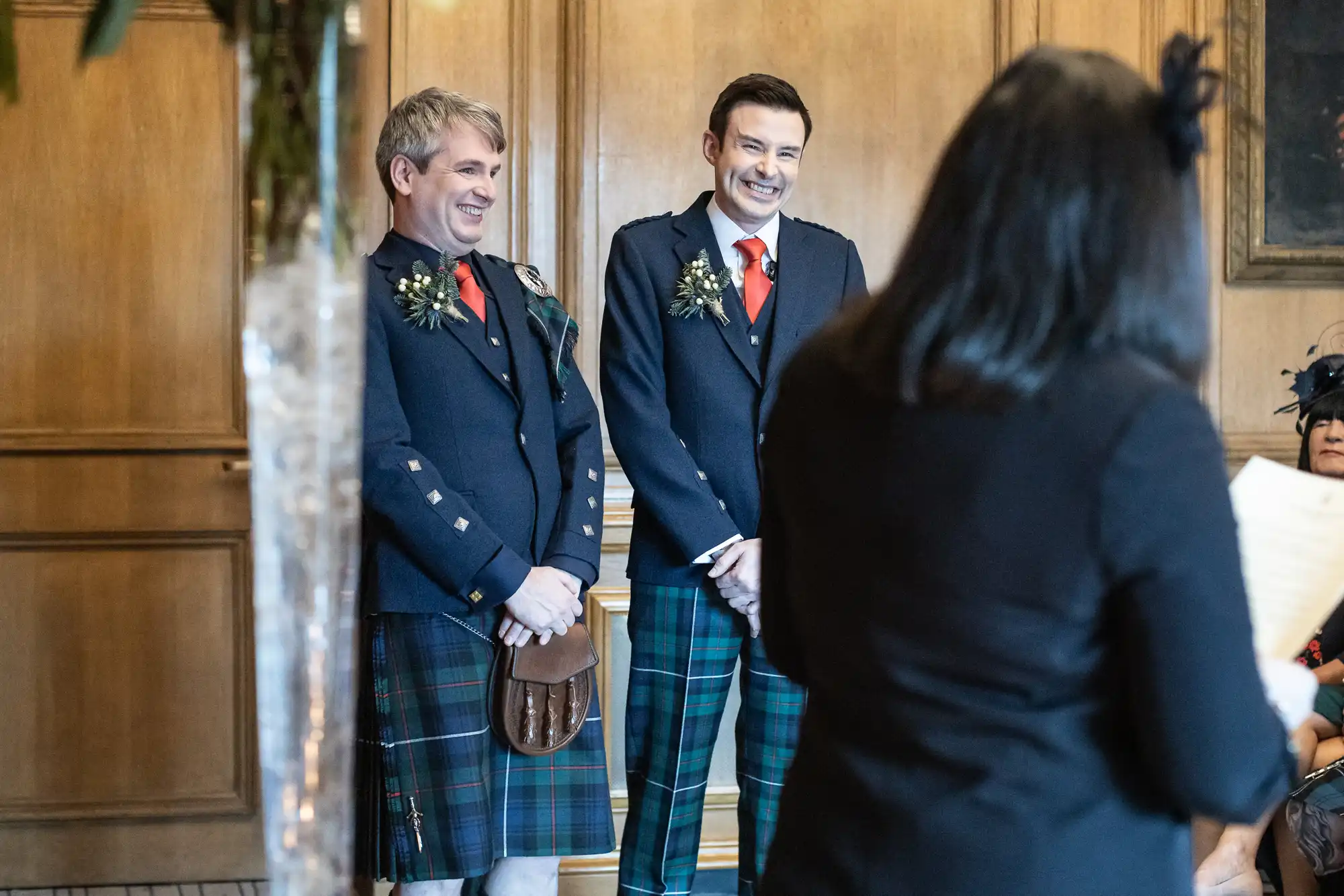 Two men in tartan kilts, decorated with floral boutonnieres, stand smiling in a room with wooden paneling. A woman in the foreground has her back to the camera and appears to be speaking.